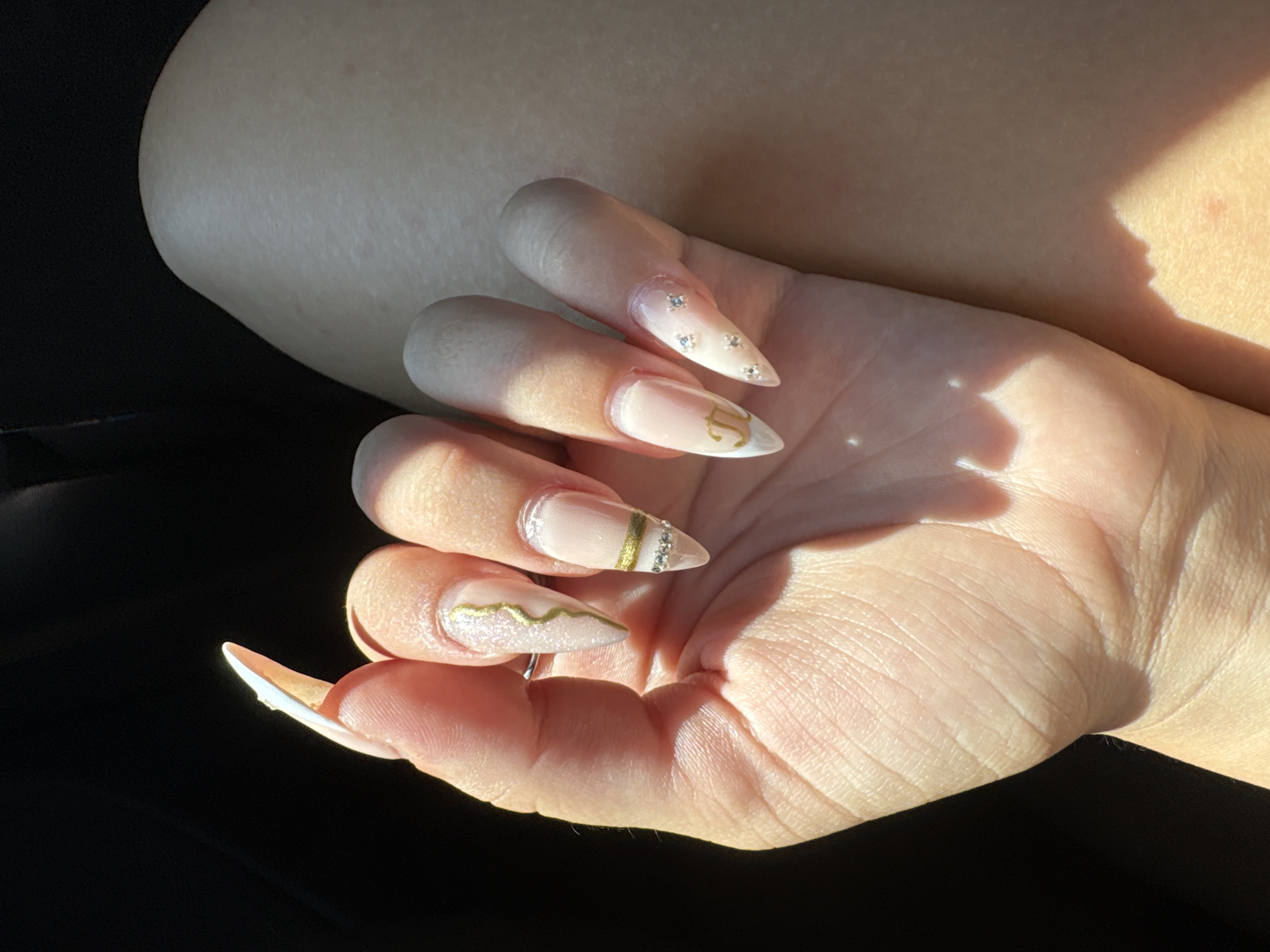 In an outdoor, sunlit environment, a person's hand is positioned against a dark background and a lightly textured, pale surface. The hand displays a set of stiletto-shaped, acrylic nails, each adorned with a unique design. The thumb nail features a swirled gold stripe and subtle shimmer. The index finger nail has a delicate gold line, a band of tiny rhinestones, and a French tip. The middle finger nail is decorated with a gold letter "P" and small scattered rhinestones. The ring finger nail exhibits a horizontal gold band above a row of rhinestones. The pinky nail is adorned with scattered small rhinestones. The skin on the hand shows natural creases and texture, with sunlight casting distinct shadows. The pale surface next to the hand is likely clothing or skin, showing a soft texture and slight variation in tone due to lighting.