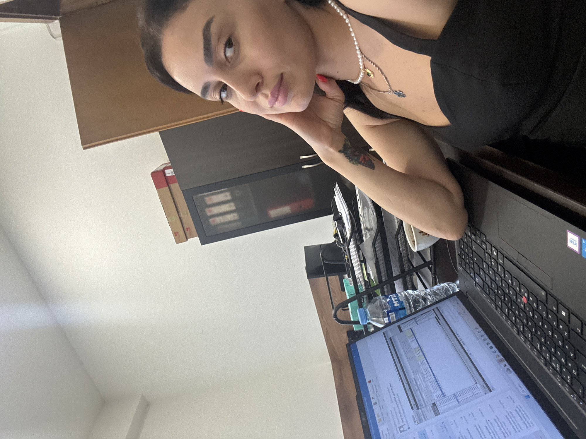 The image shows a woman sitting at a desk in what appears to be an office environment. She is looking at the camera and resting her head on her hand. On the desk in front of her is a laptop, a bottle of water, and a cup. In the background, there are shelves with folders and boxes. The overall impression is of a person working in an office, possibly taking a break or posing for a photo. There is no visible indication that this location is in Tbilisi, Georgia.