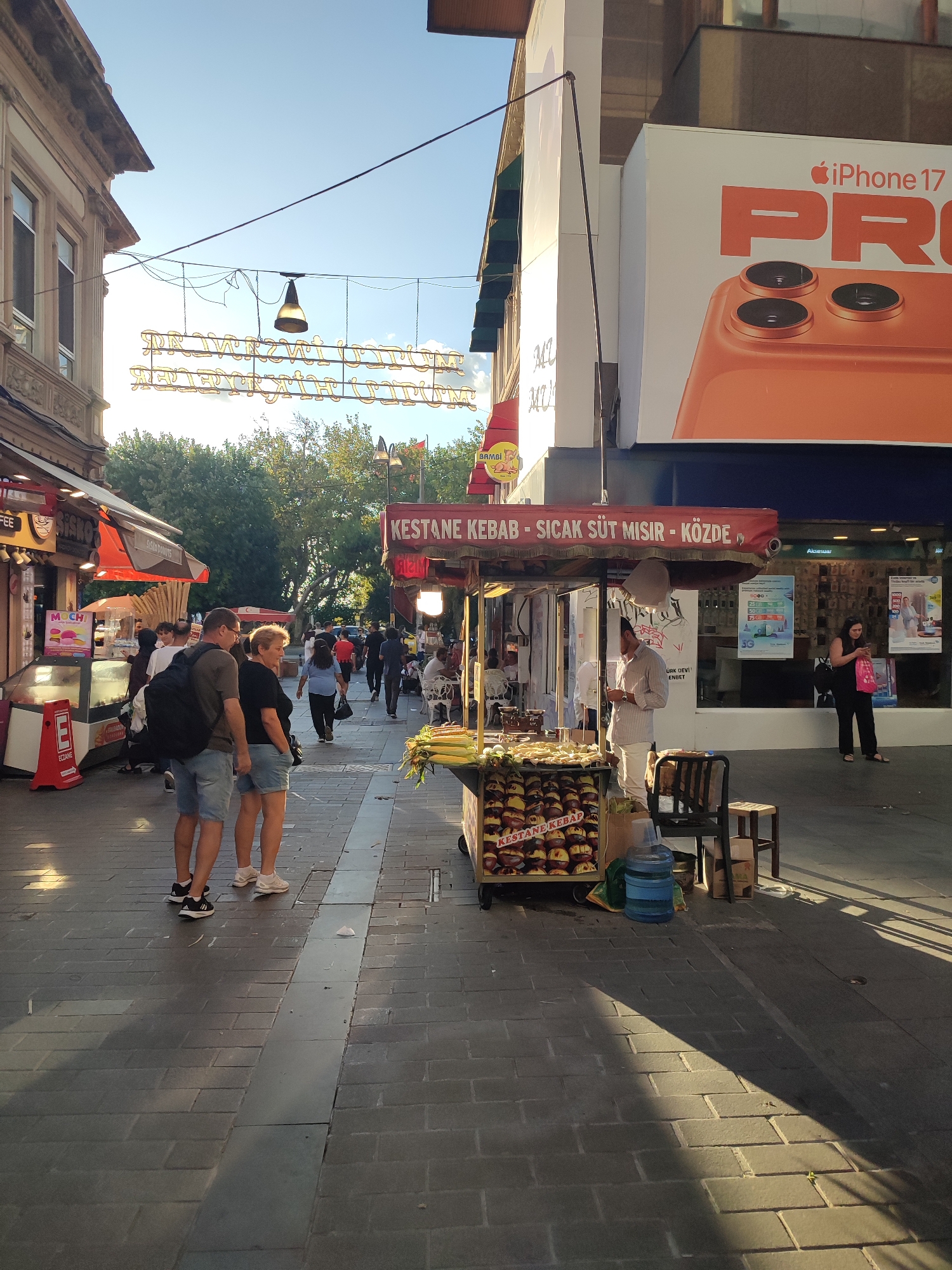 The image captures a street scene in Kadıköy, Turkey, bathed in the warm glow of late afternoon sun. The foreground is dominated by a pedestrian walkway paved with dark grey interlocking bricks, with a metallic strip running down the center. To the right, a food cart is laden with roasted chestnuts labeled "KESTANE KEBAP," and stacks of corn on the cob. Two people, a man and a woman, stand facing away from the camera, their backs towards the viewer, appearing to be a couple. The man wears a dark grey t-shirt and blue shorts, carrying a black backpack. The woman is in a black t-shirt and denim shorts.
Further down the street, more people are visible, some walking, others browsing at stalls. To the left, there are shops with awnings, one displaying "MOCHI" and another "BIŞKO." Overhead, decorative lights with a neon glow are strung across the street. A large billboard advertising "iPhone 17 PRO" is prominent on the right side of the street. The environment suggests a busy commercial area with a mix of food vendors and retail establishments. The shadows cast by the setting sun stretch across the pavement, adding depth and ambiance to the scene.