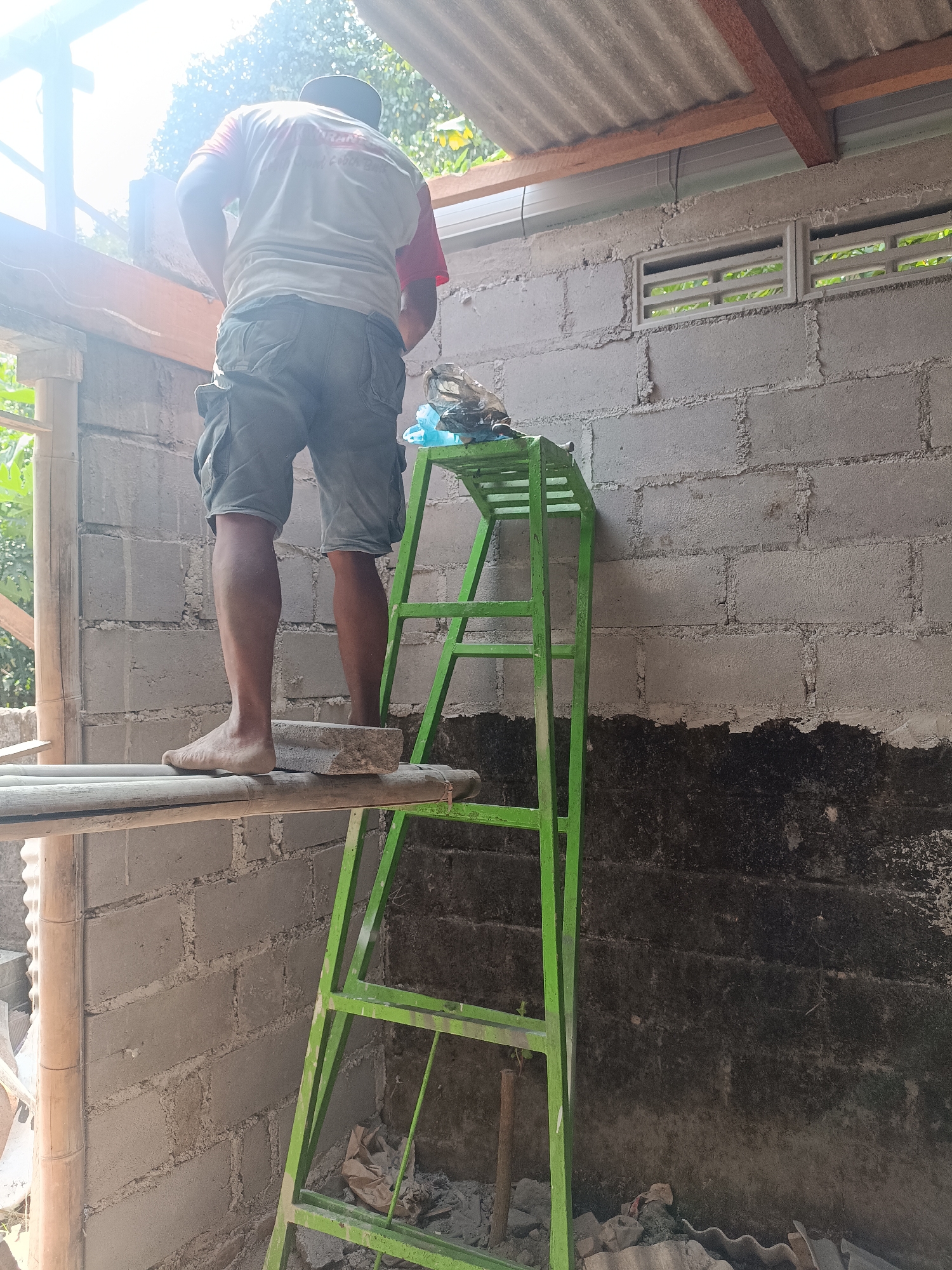 In the city of Sumberadi, Indonesia, a construction worker stands on a wooden platform, balancing on a brick. He is wearing a white t-shirt with red sleeves and grey cargo shorts. In front of him is a green metal ladder with a few items on its top shelf. The worker is positioned near a grey concrete block wall, part of which is covered in dark, tar-like material. Above the wall are two rectangular vents. The scene appears to be an unfinished building, with corrugated metal roofing visible overhead. The overall atmosphere is one of ongoing construction and manual labor.