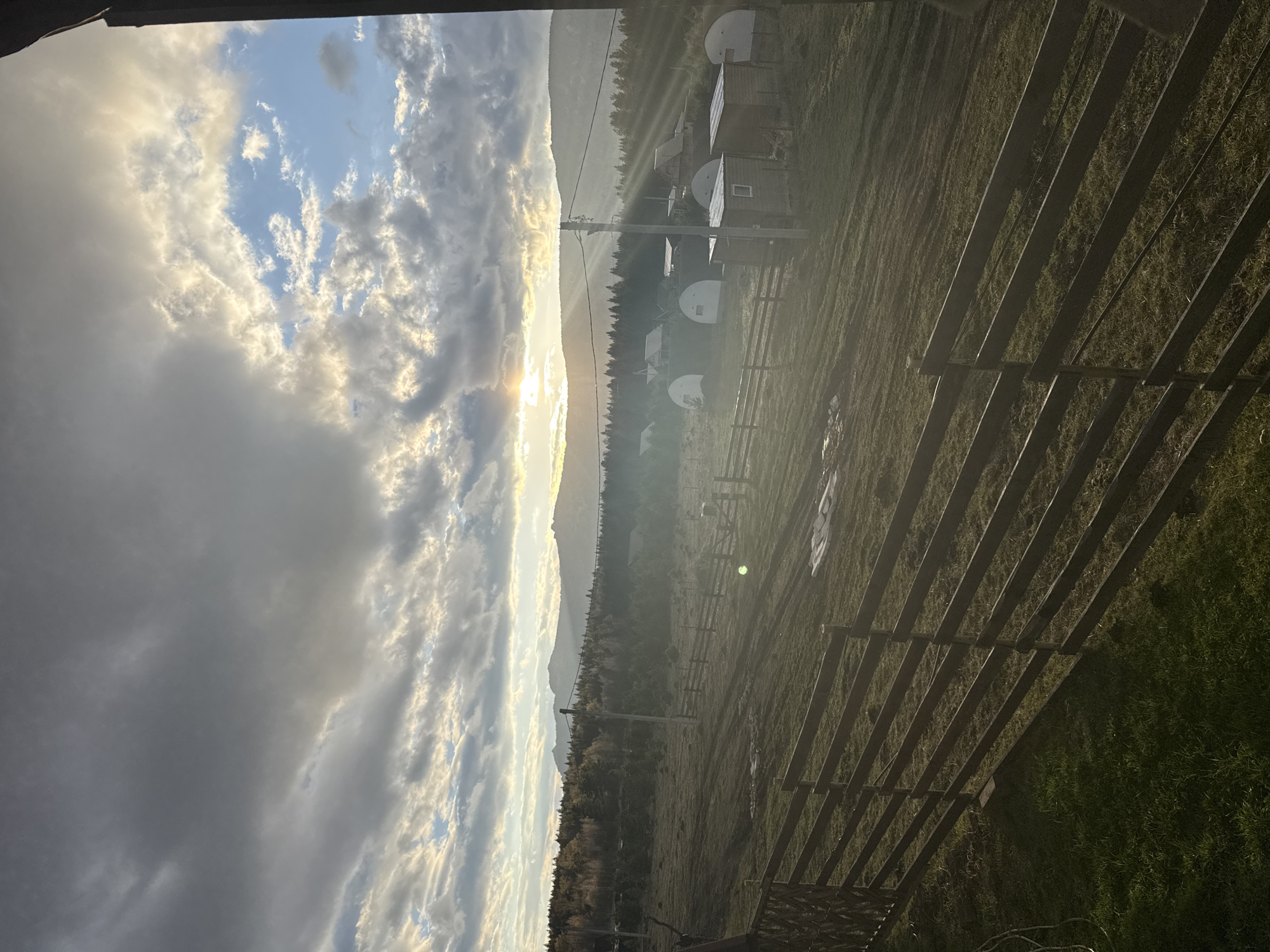 The image shows a landscape view under a sky with clouds and a bright sun. In the mid-ground, a line of trees separates the sky from an area with what appear to be small buildings with rounded roofs. A wooden fence spans the foreground, partially obscuring a grassy area. The location is likely in the vicinity of Tyulyuk, Russia, but further location information is not possible without additional information.