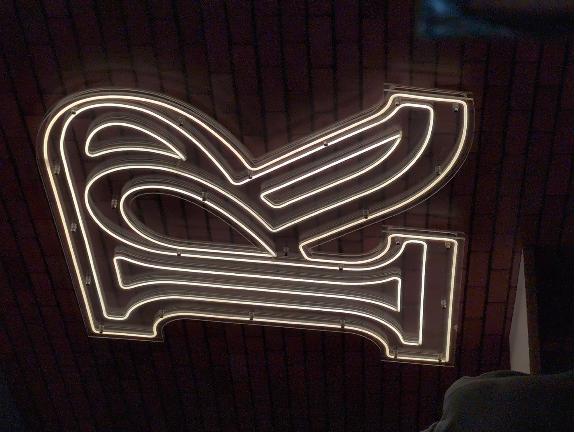The image depicts an illuminated neon sign shaped like a stylized Russian "R" (Р) character against a dark brick wall background. The neon light is a warm white color. The sign is mounted on the wall with visible metal standoffs. A person wearing a grey or green shirt is visible in the lower right corner of the image, partially obscured. The location is likely Izhevsk, Russia, as the sign appears to be for the "Rossiya" hotel.