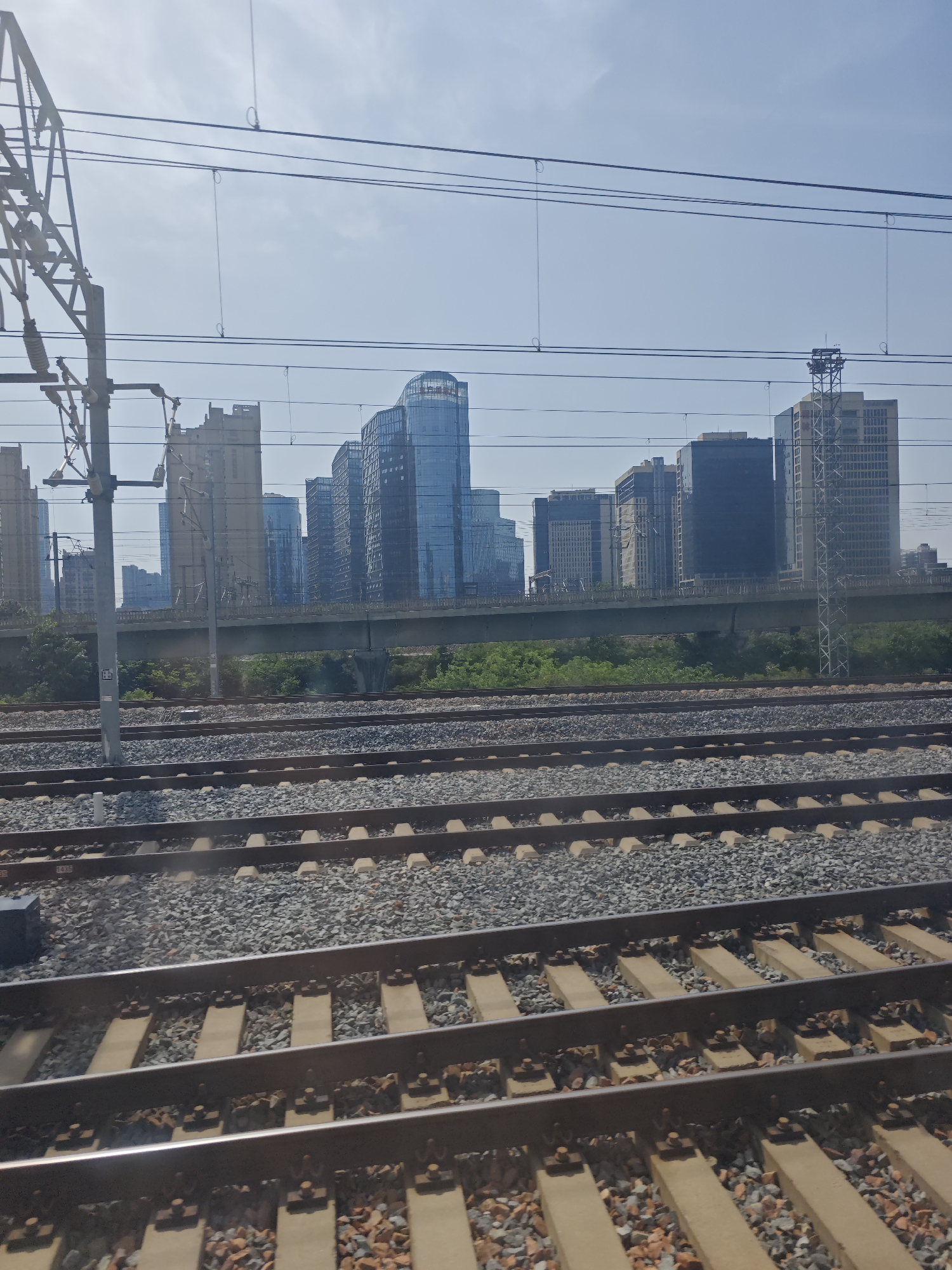 A daytime photograph captures a panoramic view from alongside multiple train tracks towards a modern urban skyline under a clear, bright sky.
In the foreground, several parallel sets of railway tracks stretch into the distance. The tracks are laid on grey and reddish ballast, with concrete sleepers holding the metal rails in place. To the left, a tall metal structure, part of an overhead catenary system for electric trains, rises, supporting numerous power lines that crisscross the upper part of the frame. A small white tag with indistinct black text is visible on the base of this pole. On one of the concrete sleepers in the lower left, the number "1435" can be faintly discerned.
Beyond the tracks, a strip of green vegetation is visible, followed by a wide concrete bridge or overpass that spans horizontally across the scene. In the background, a dense cluster of modern high-rise buildings forms a city skyline. Many of these skyscrapers feature reflective blue-tinted glass facades, while others are built with more traditional concrete and window designs. One prominent building on the far right has a lattice-like structure on its side. A faint white logo or text is visible on the top of one of the blue-glass buildings, but it is unreadable. The overall setting suggests a bustling urban or peri-urban environment, likely viewed from a stationary train or a platform beside the railway line on a sunny day.