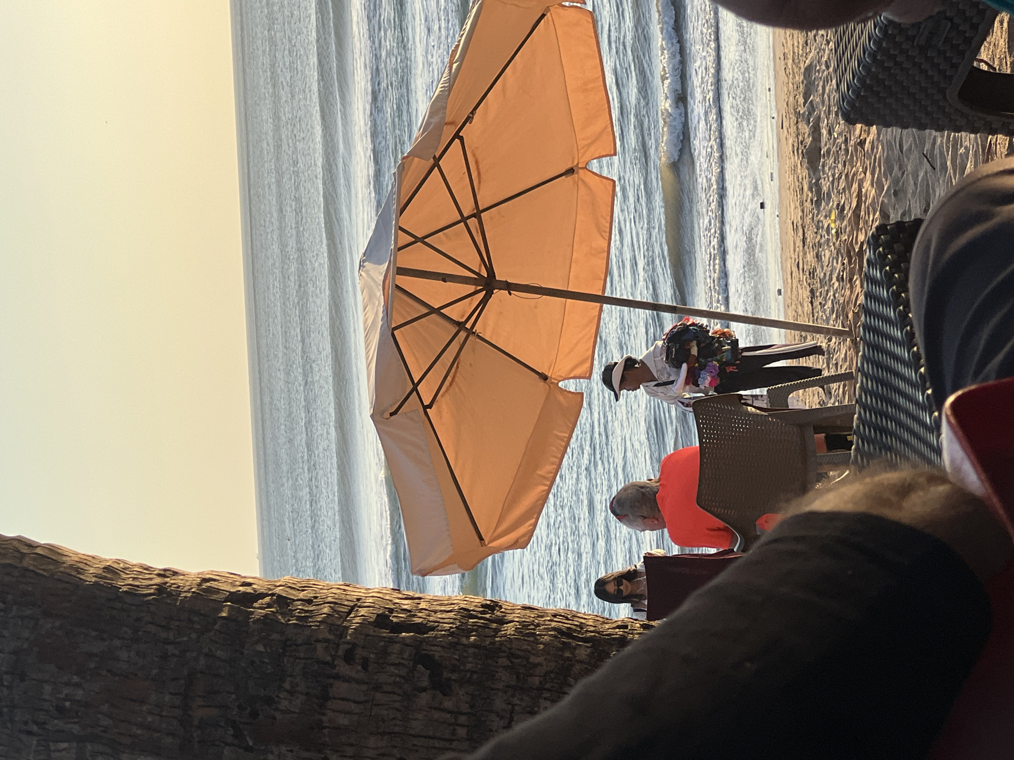 The image depicts a beach scene at dusk, likely in Kuta, Indonesia. The foreground is dominated by the dark silhouette of a palm tree trunk on the left and parts of outdoor seating furniture, suggesting a beachside cafe or restaurant. In the midground, an orange beach umbrella casts a shadow over the sandy beach. Several individuals are visible. Two people are seated, one in an orange shirt facing away from the camera, and another person, possibly a woman, seated to their left, wearing sunglasses and looking down at an object. A third person, standing near the umbrella and appearing to be a vendor, holds a collection of colorful items. The background features the ocean with gentle waves and a hazy, pale yellow sky, characteristic of sunset. The overall atmosphere is relaxed and tropical.