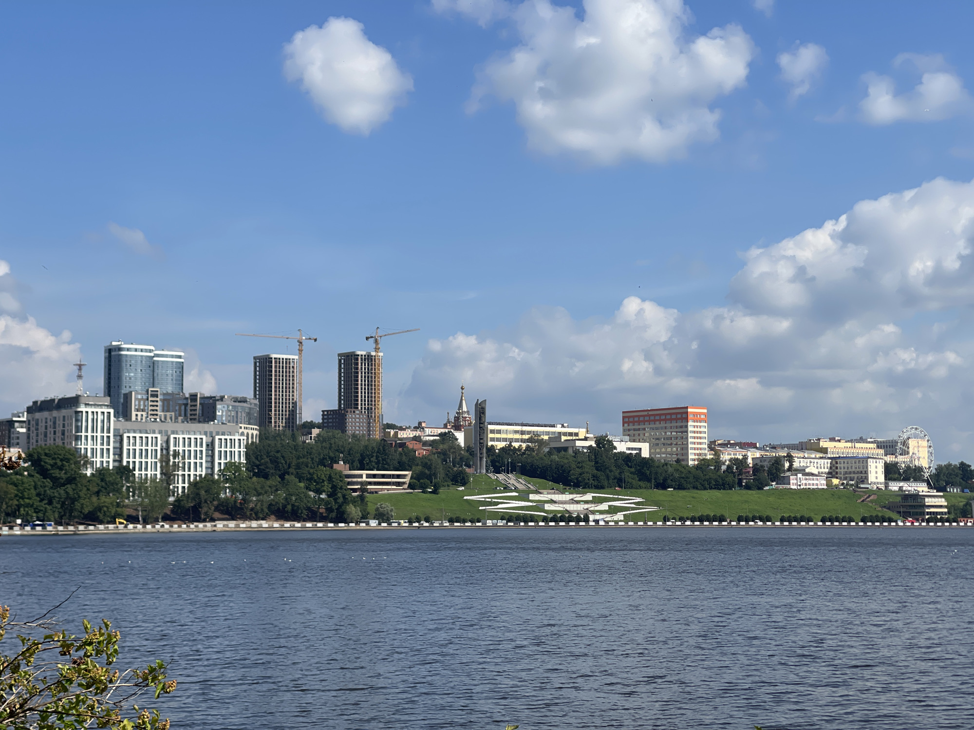 The image depicts a wide, blue river in the foreground, with a city skyline visible across the water on a sunny day with scattered white clouds in a bright blue sky. The city features a mix of modern high-rise buildings, older structures, and greenery on a sloping embankment. Prominent structures include several tall, contemporary residential buildings with glass facades, some under construction with cranes visible. There are also low-rise apartment buildings, and a few older, multi-story buildings, one with an orange and white facade. A large monument with a tall obelisk and surrounding tiered grassy area with a decorative pattern is situated on the embankment, with a domed church spire visible behind it. Further to the right, a Ferris wheel stands against the skyline. The riverbank is lined with trees and a white and black decorative border. The water shows ripples, and a few small white birds are visible on its surface. In the lower left corner, a small portion of a green leafy branch with seed pods is in focus. The overall impression is of an urban waterfront with both new development and established architecture. This scene is likely the Izhevsk Pond (or Izhevsk Reservoir) in Izhevsk, Russia, given the prominent Ferris wheel and the general appearance of the city's waterfront.