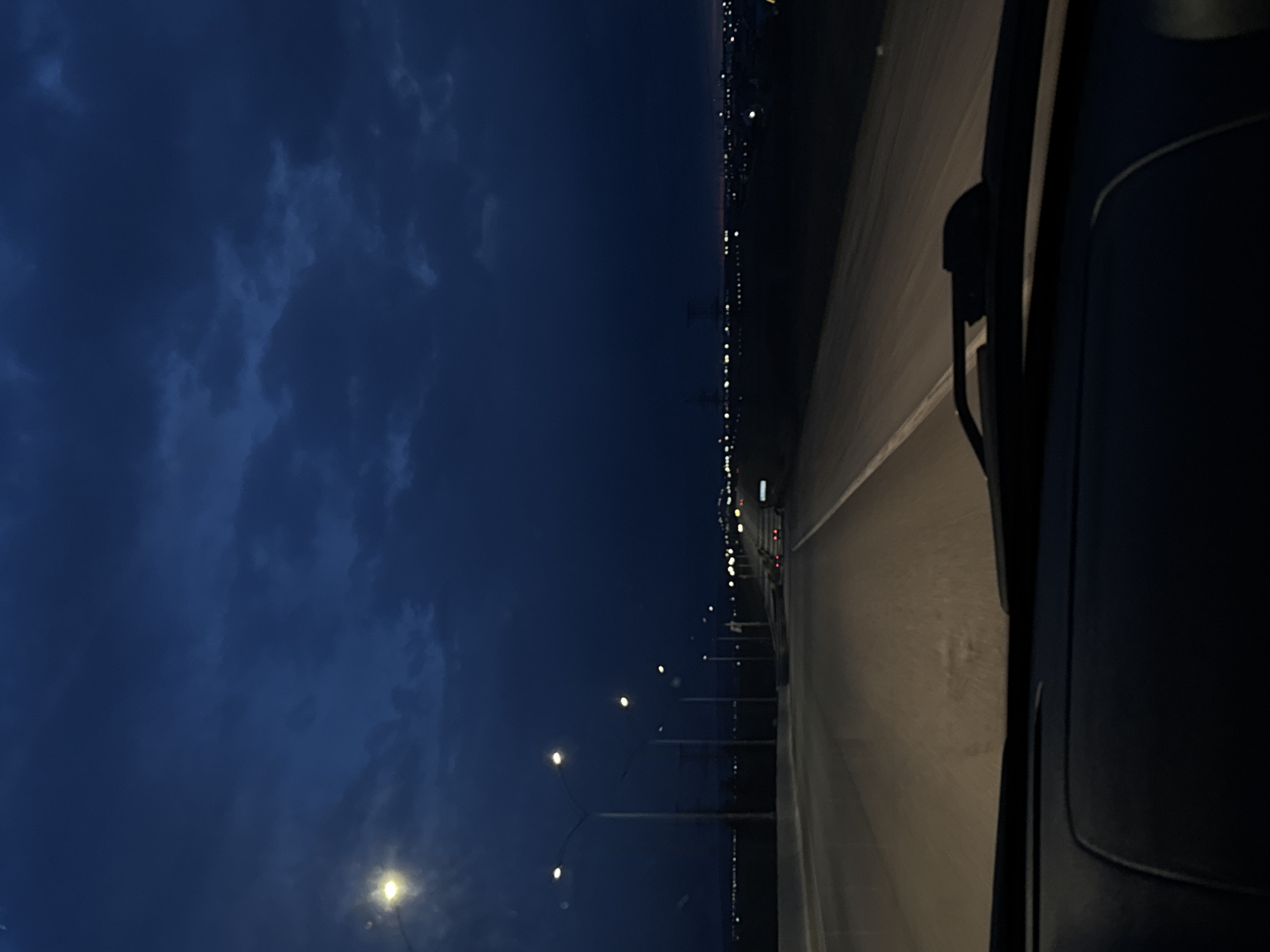 The image captures a nocturnal or late twilight scene from the perspective of a moving vehicle, looking towards the front passenger side. The scene is dominated by a dark blue sky with indistinct, shadowy clouds occupying the upper left quadrant of the frame.
A long, straight stretch of road, possibly a highway or bridge, runs horizontally across the middle and right side of the image, appearing to recede into the distance. The road surface is dimly lit, likely by streetlights and the vehicle's headlights, with reflections of light visible on its surface. Along the edge of the road, a series of streetlights with bright, circular glows extend into the distance, illuminating the immediate area. In the far distance, a faint line of numerous small, bright lights suggests a city, town, or industrial area along the horizon, creating a stark contrast against the dark sky and landmass.
Part of the vehicle's interior, specifically a dark dashboard or door panel, and a windshield wiper are visible on the far right edge of the frame, confirming the vantage point from inside a car or similar vehicle. No people are visible in the image.
The time of day is clearly night or very late dusk due to the deep blue sky and the reliance on artificial lighting. The presence of clouds indicates a non-clear sky, but no other specific weather conditions like rain or fog are apparent. There is no visible text in the image. Given the context of Bol'shaya Mos', Russia, this could be a view of a road or bridge leading towards or through a settlement.