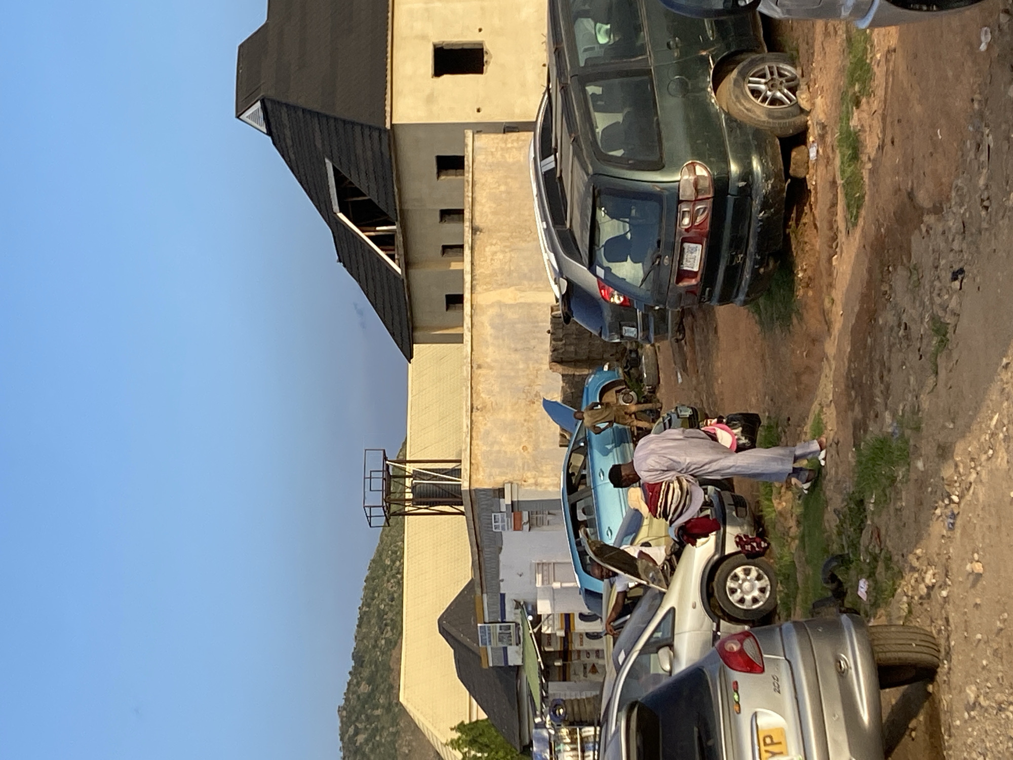 The image depicts an outdoor scene in Gwong, Nigeria, with several cars parked on a dirt and gravel area. The setting appears to be a makeshift car lot or repair area. In the background, there is a partially constructed building with a dark, sloped roof and lighter colored walls, featuring several dark, rectangular openings that suggest windows or doorways. To the left of the building, a lush, green hillside is visible under a clear, pale blue sky, indicating it is daytime. The lighting suggests it might be late afternoon or early morning, given the warm tones on the building.
In the foreground, a group of people are interacting around a light blue car. One person, dressed in a light gray robe and headscarf, is leaning over the open hood of the car, appearing to be working on it or examining something inside. Another individual, wearing a patterned headwrap and a maroon top, is also near the car. Several other vehicles are parked in the vicinity, including a dark green SUV and a silver hatchback. One of the silver cars has a visible license plate that reads "YP". The overall atmosphere seems busy and functional, possibly a place where car repairs or sales take place.