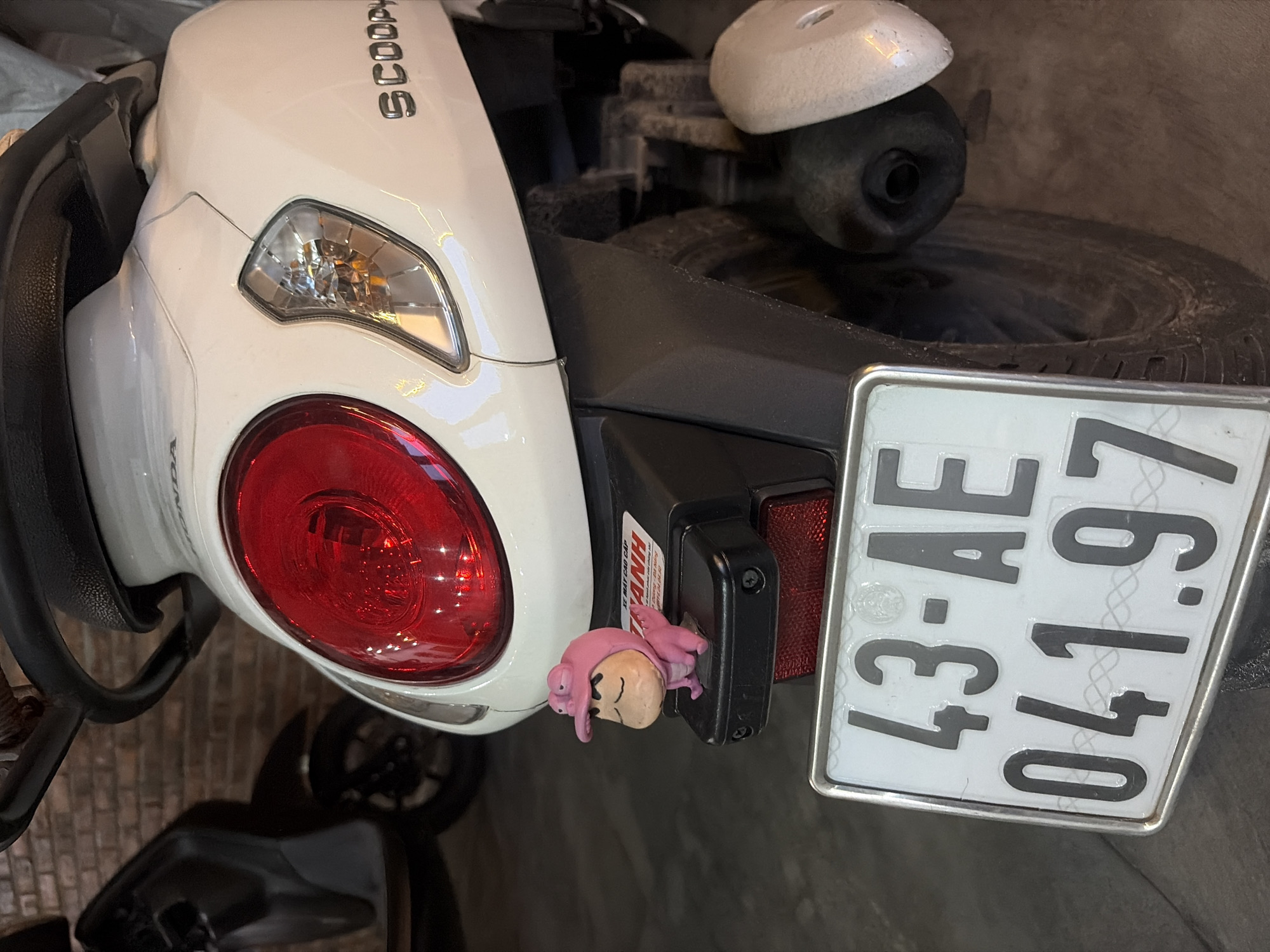 The image shows the rear of a white Honda Scoopy scooter parked outdoors, with a brick wall visible in the background. A black scooter is partially visible to the left. The license plate of the Honda Scoopy reads "43-AE 041.97". A small figurine of a character wearing a pink dinosaur costume is attached to the rear fender, positioned above the license plate. The scooter's red taillight and clear turn signal are prominent features. The ground appears to be a concrete or asphalt surface. The presence of the "43" prefix on the license plate indicates that the scooter is registered in Đà Nẵng, Vietnam.