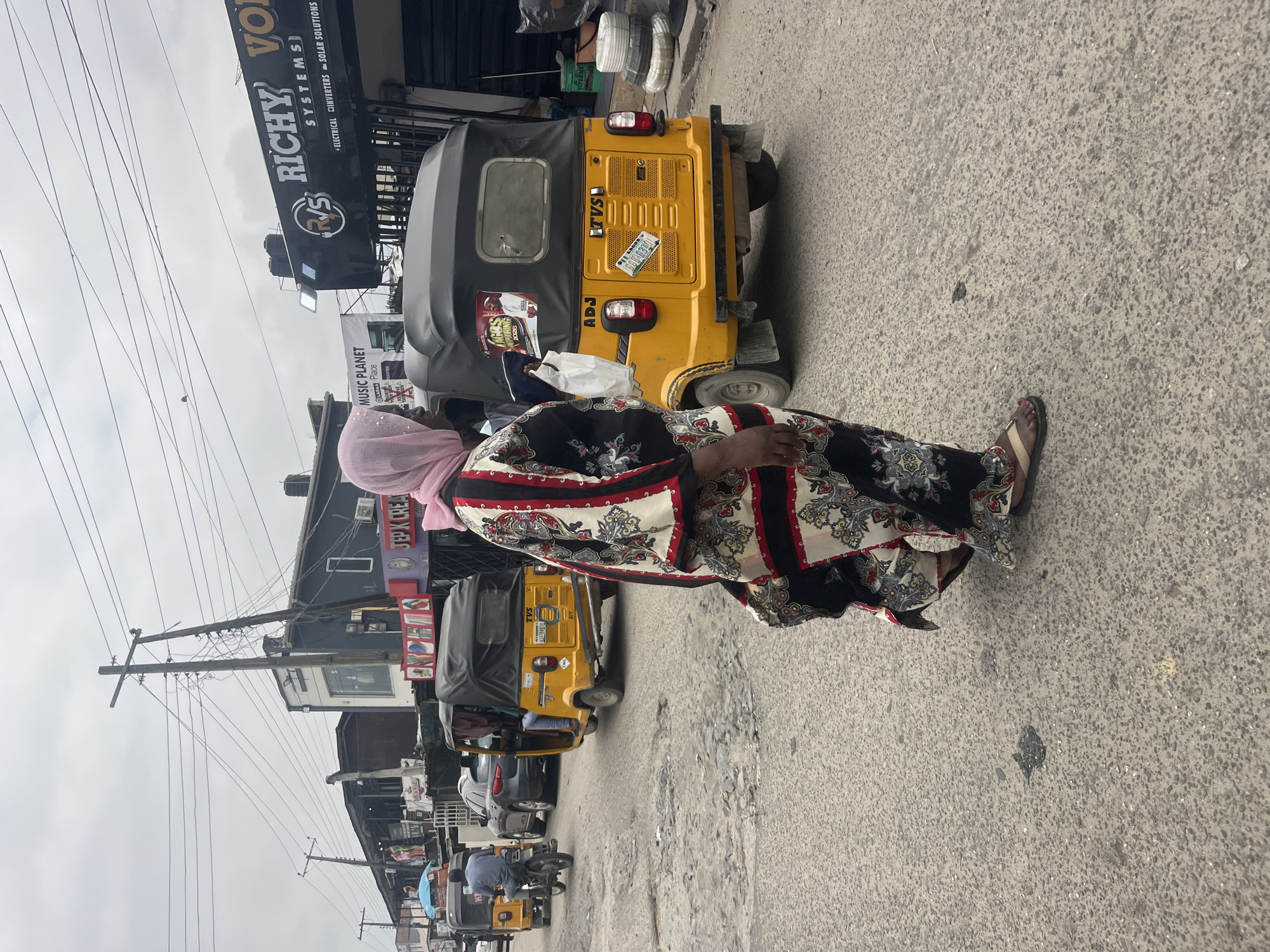 In Ado, Nigeria, a woman wearing a pink hijab and a black and white patterned boubou walks on a street with a rough, patchy asphalt surface. She is the primary subject in the foreground, with her back mostly to the camera. Behind her, several yellow motorized tricycles, commonly known as "Keke NAPEPs" or "Marwas" in Nigeria, are present on the road. One tricycle is in motion, driven by a person whose legs are visible. Another tricycle is stationary. The street is lined with various businesses, including shops with signage such as "RICHY VOL SYSTEMS" and "JEP K CREA." Electrical wires are strung overhead, supported by wooden utility poles. The sky is overcast.