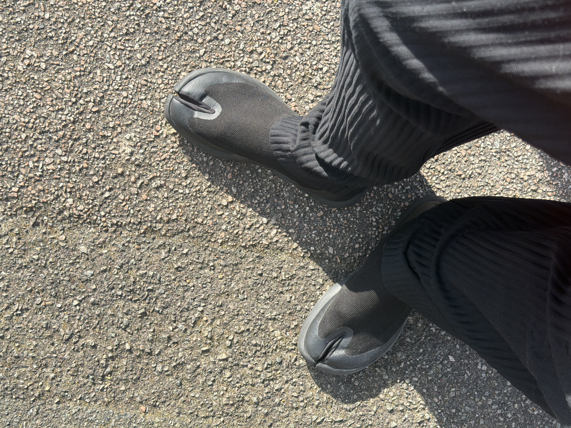 An overhead shot depicts a person wearing black split-toe shoes standing on asphalt. The shoes appear to be a sock-like material extending up the ankle, and the person is also wearing black, pleated trousers. The asphalt is typical of roadways or pavements, made of small stones and gravel set in a binding agent. The location is likely a paved surface within the Folkestone and Hythe District in the United Kingdom, given the type of asphalt used. There are no other people visible in the frame. The action being depicted is the act of standing on an asphalt surface.