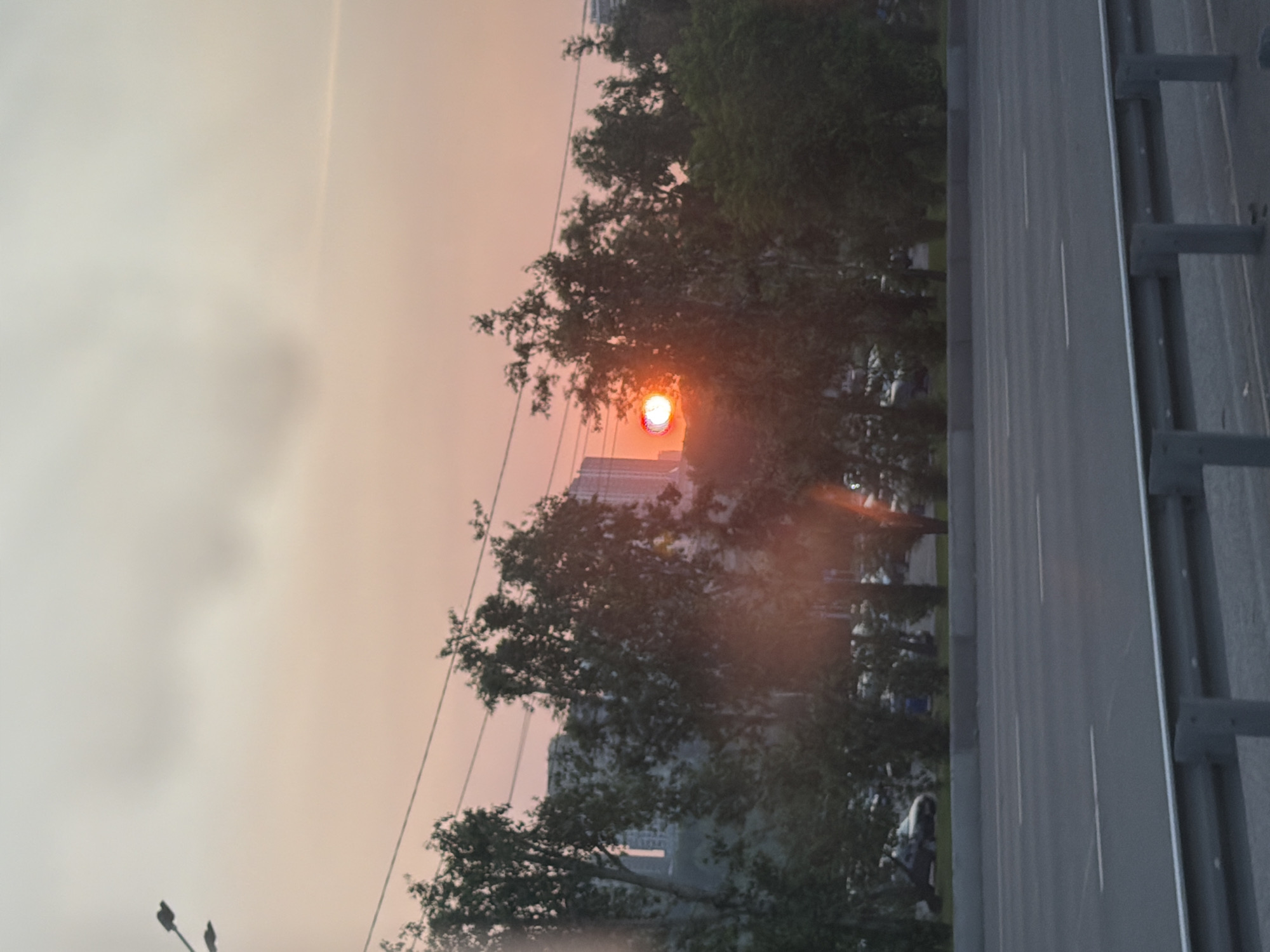 The image depicts a street scene in Krasnoyarsk, Russia, during what appears to be sunset. The sky is a hazy gradient of orange and pink, suggesting atmospheric conditions that could be due to dust or pollution, common in industrial areas. A bright, reddish-orange orb, likely the sun, is visible through the foliage of trees, partially obscured and appearing diffused.
On the left side of the frame, silhouettes of trees are prominent, with some branches reaching across the scene. In the background, the indistinct shapes of buildings can be seen, suggesting an urban or semi-urban environment. The presence of streetlights on poles indicates an infrastructure consistent with a city.
The right side of the image is dominated by a paved road, possibly a highway or a wide city street, with clear lane markings. A concrete barrier with metal railings runs along the side of the road, typical of elevated roadways or bridges. The perspective is from above, looking down at the road.
While the image is largely devoid of sharp details, there are hints of activity. A few figures can be discerned among the trees, and a vehicle might be present on the road, though not clearly identifiable. The overall mood is somewhat muted, likely due to the hazy lighting and the overcast sky, contributing to a sense of quietude rather than vibrant activity. The time of day is clearly indicated by the sunset colors in the sky and the diffused sunlight. No visible text is present in the image.