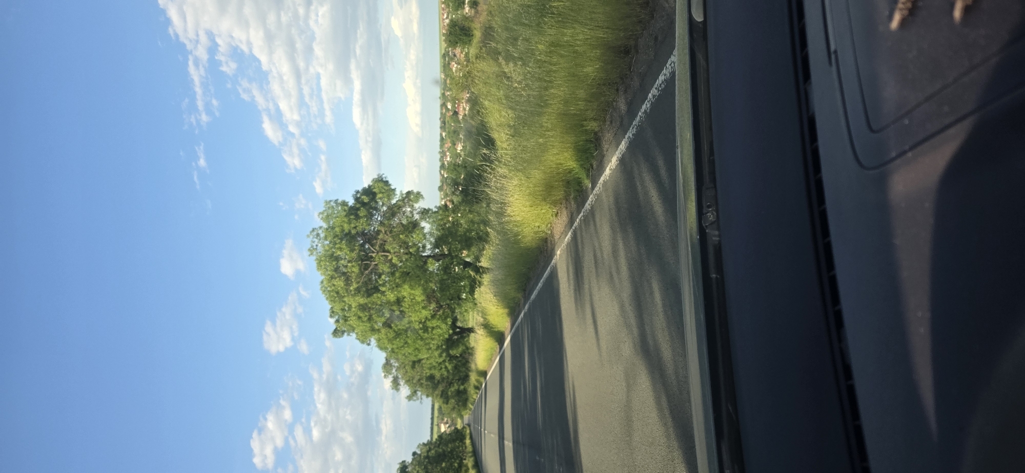 The image depicts a view from inside a moving vehicle on a road near Troyanovo, Bulgaria. The road is asphalt and has a white line marking the edge. Vegetation, including tall grass and trees, lines the roadside. In the distance, glimpses of buildings suggest a town or village. The sky is blue with scattered clouds. Part of the vehicle's interior is visible in the foreground, including a dark dashboard.