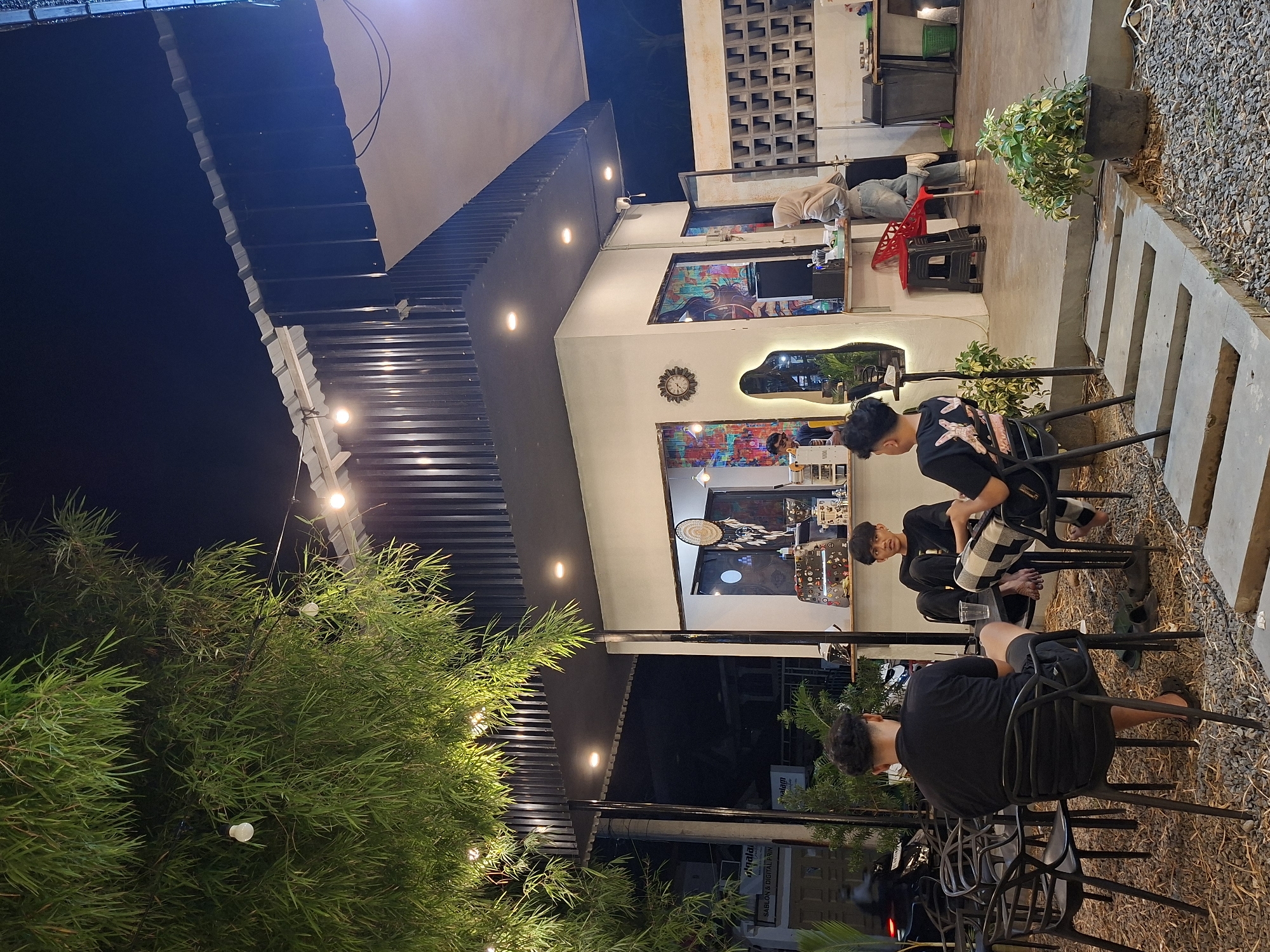 The image depicts an outdoor seating area, presumably of a cafe or restaurant, in Lhokseumawe, Indonesia. Three individuals are visible, seated on chairs. The setting is at night, as indicated by the dark sky. The architecture features a blend of modern design, with clean lines and contemporary lighting. There are decorative elements, including wall art and plants. Streetlights and the cafe's lighting provide illumination. The ground is covered in gravel around the seating area.