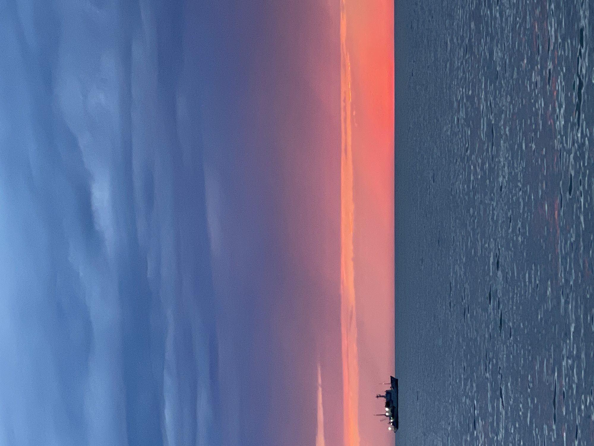The image depicts a sunset scene in what appears to be a coastal environment, possibly near Sochi, Russia. A body of water, likely the Black Sea, occupies the right side of the frame, with a subtle reflection of light indicating a slightly disturbed surface. A ship or boat is visible on the water's surface, near the horizon. The sky transitions from a deep blue on the left to shades of purple and then a vibrant orange and pink near the horizon line. Clouds are present in the sky, contributing to the layered effect of the sunset. The image is oriented vertically.