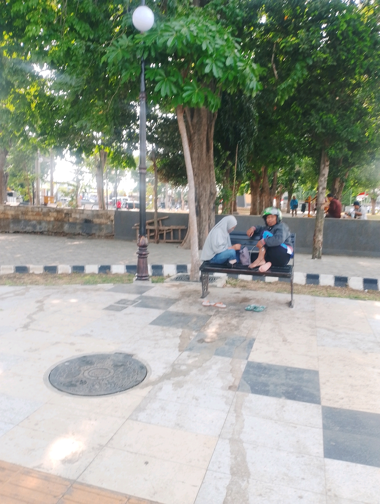 The image depicts an outdoor scene in Probolinggo, Indonesia, likely a park or public square. In the foreground, a paved walkway with a checkered pattern of light and dark tiles leads to a circular manhole cover. A lamp post with a round light fixture stands near the center of the image, surrounded by trees with lush green foliage.
In the midground, two individuals are seated on a black metal bench. One person, wearing a light grey hijab and blue jeans, is looking down at something in their lap. The other person, wearing a dark jacket, black pants, and a green helmet, is looking towards the first person. Both are barefoot. Nearby, on the ground, are a pair of light-colored sandals and a pair of green flip-flops. The bench is situated on a paved area bordered by a black and white striped curb.
The background reveals more trees, a low stone wall, and glimpses of buildings and other people in the distance, suggesting a populated area within the city. The overall atmosphere appears to be relaxed and casual, with the subjects enjoying a moment of rest outdoors.