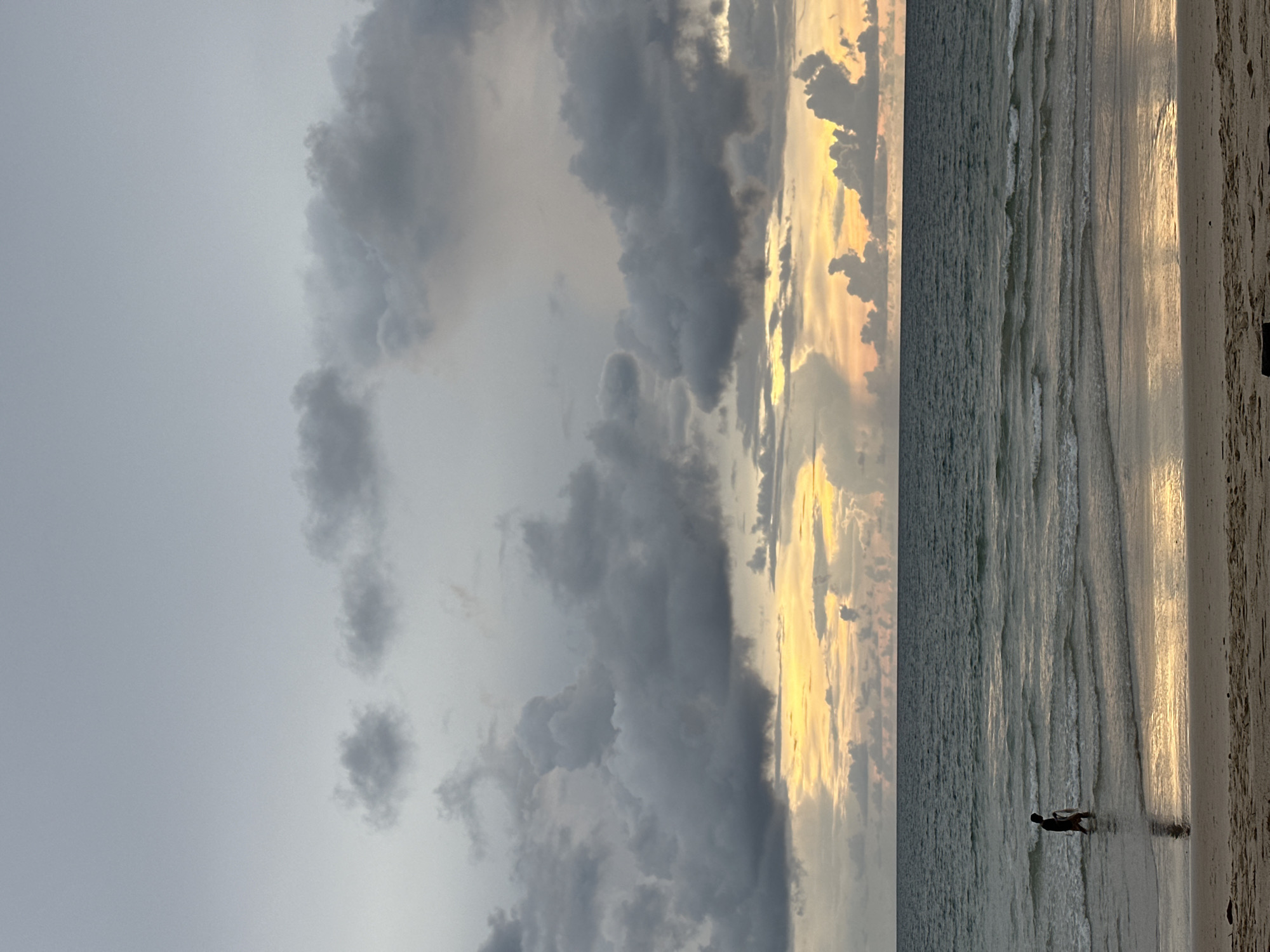 A silhouette of a lone person stands in the shallow water of a beach. The person is facing away from the camera and appears to be holding something in their hand. The water is calm with gentle waves lapping at the shore, reflecting the warm hues of the sunset sky. The sky is filled with dramatic, dark clouds illuminated by streaks of orange and yellow from the setting sun. The sand in the foreground is light brown. The scene depicts a tranquil, somewhat moody atmosphere at dusk on an unspecified beach.
