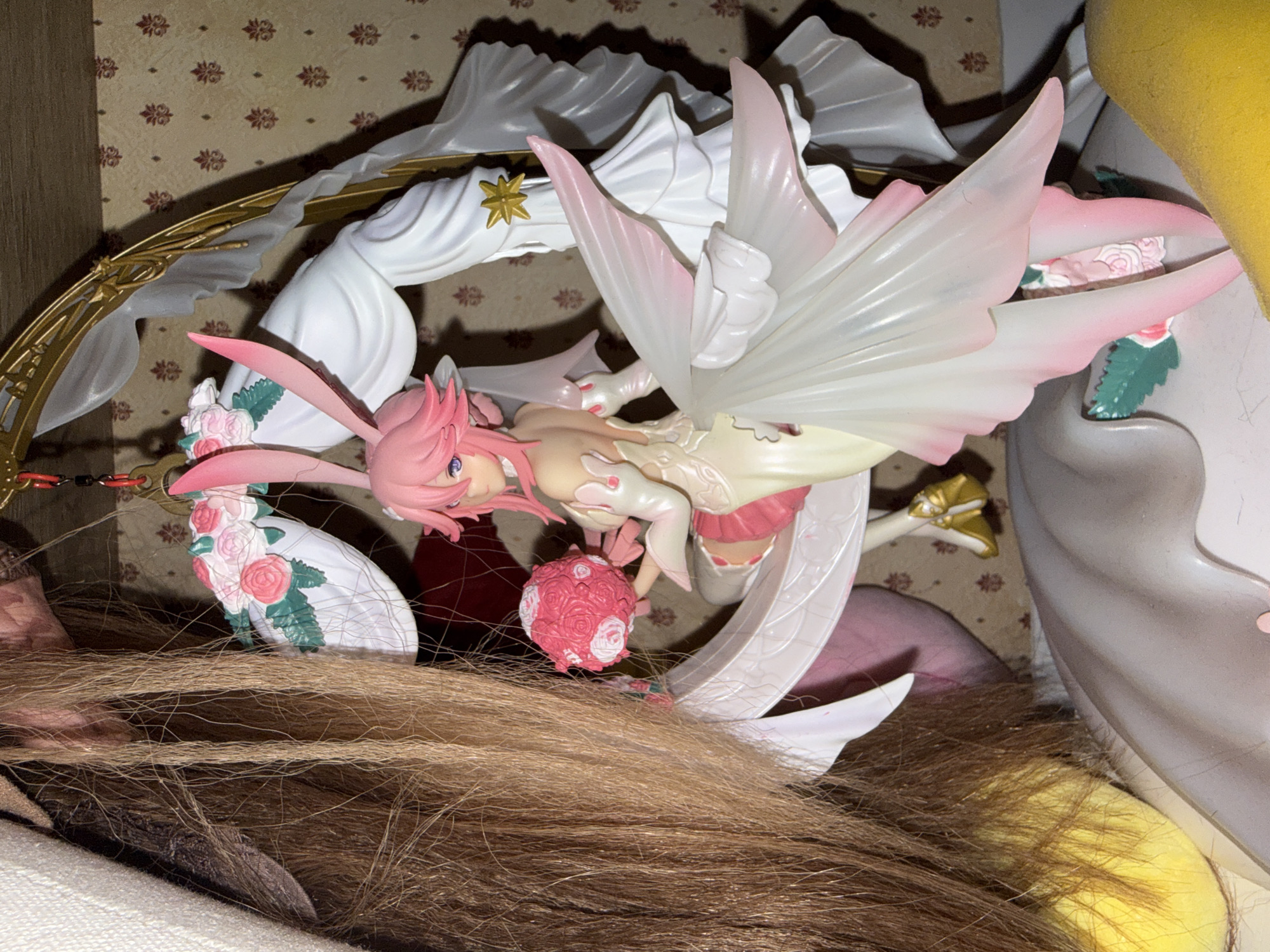 The image shows a close-up of a figure of a female character with pink hair and bunny ears, wings, and a flowing white dress. The figure is surrounded by elements of its base, including a golden arc, white decorative pieces, and pink floral accents. The figure is placed on what appears to be a brown, possibly wig-like, material. The background features a patterned wall, possibly wallpaper. The image lacks specific location information within Moscow, Russia, or any human presence, limiting forensic detail.
