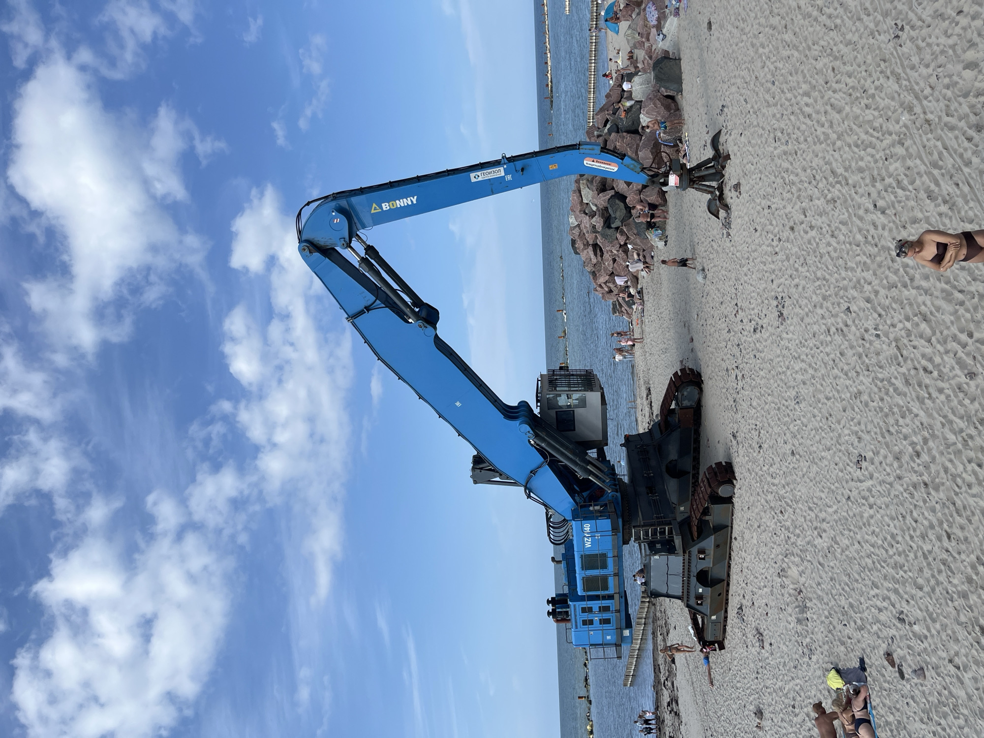 In Svetlogorsk, Russia, a large blue industrial excavator is positioned on a sandy beach next to the Baltic Sea. The excavator, a Bonny WZ140, has a long boom and is mounted on a tracked base. In the background, the sea stretches to the horizon under a sky with scattered white clouds. A breakwater constructed from large rocks is visible in the water, and on the beach, several people are sunbathing and relaxing. Some are near the excavator, while others are further down the shore, some with beach umbrellas and tents. The beach has scattered rocks and debris.