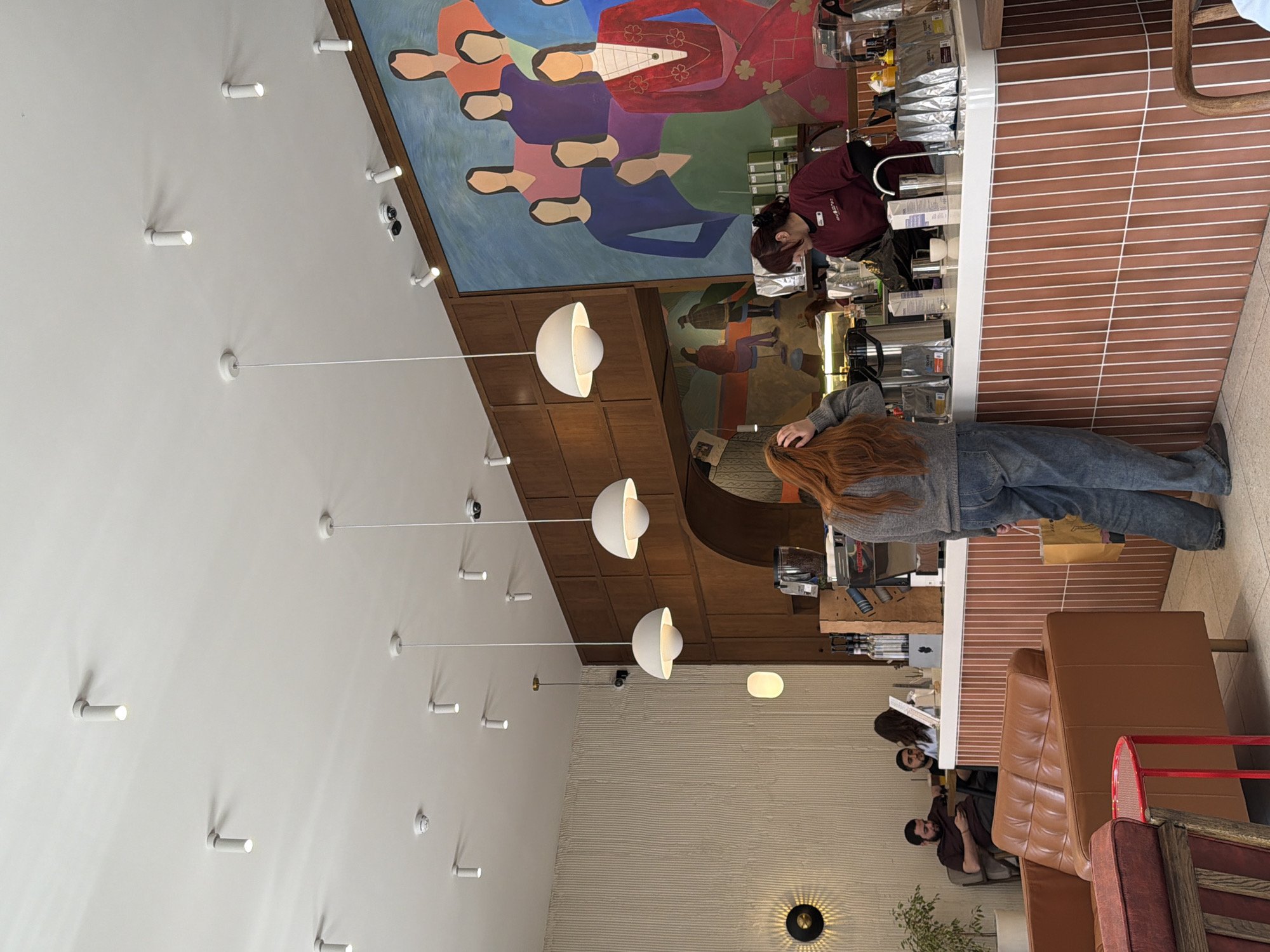 The image shows the interior of a café, possibly in Zamankul, Russia.
**People:** A barista stands behind a counter, facing right and attending to equipment. A customer with long, red hair stands at the counter, facing away from the camera. Further into the café, two seated patrons appear to be conversing.
**Subjects:** The focus is on the café's interior design.
**Objects:** A counter with coffee machines and supplies dominates the foreground. Hanging globe-shaped lights provide illumination. Artwork adorns the walls, including a large mural of figures in traditional dress above the counter, and a painting behind the customer. Seating consists of leather sofas.
**Actions:** The barista is likely preparing a beverage. The customer stands at the counter, possibly placing an order. Seated patrons are conversing.
**Interactions:** A transaction between the customer and barista is implied. The patrons are interacting in conversation.
**Environment:** The café features a modern, eclectic design. Wood paneling and a textured wall provide architectural details. Track lighting is visible on the ceiling. The flooring consists of brown and white stripes.