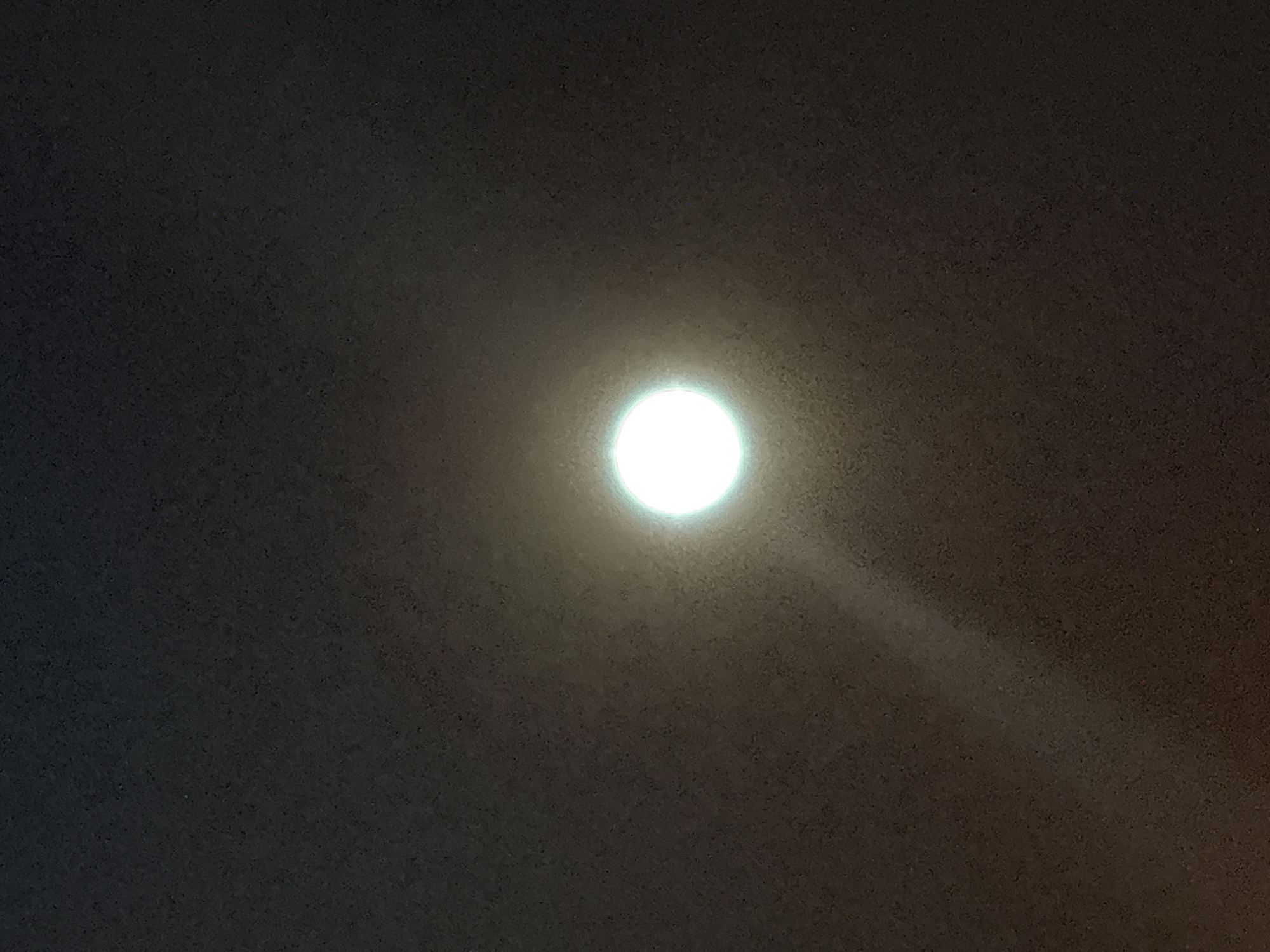 The image shows a bright, circular light source against a dark, textured background. The light source appears to be a bulb or a similar fixture, emitting a strong white glow with a subtle green halo. A faint beam of light emanates from the bottom right of the light source, extending diagonally downwards and to the right, illuminating the textured surface. The background texture is a speckled, dark gray or black, resembling a ceiling or wall with a rough finish. The overall impression is of artificial lighting in a dimly lit indoor environment. The image does not provide any information about the city of Sveti Vlas, Bulgaria, or any specific location within it. There are no people, objects, or actions depicted in the image.