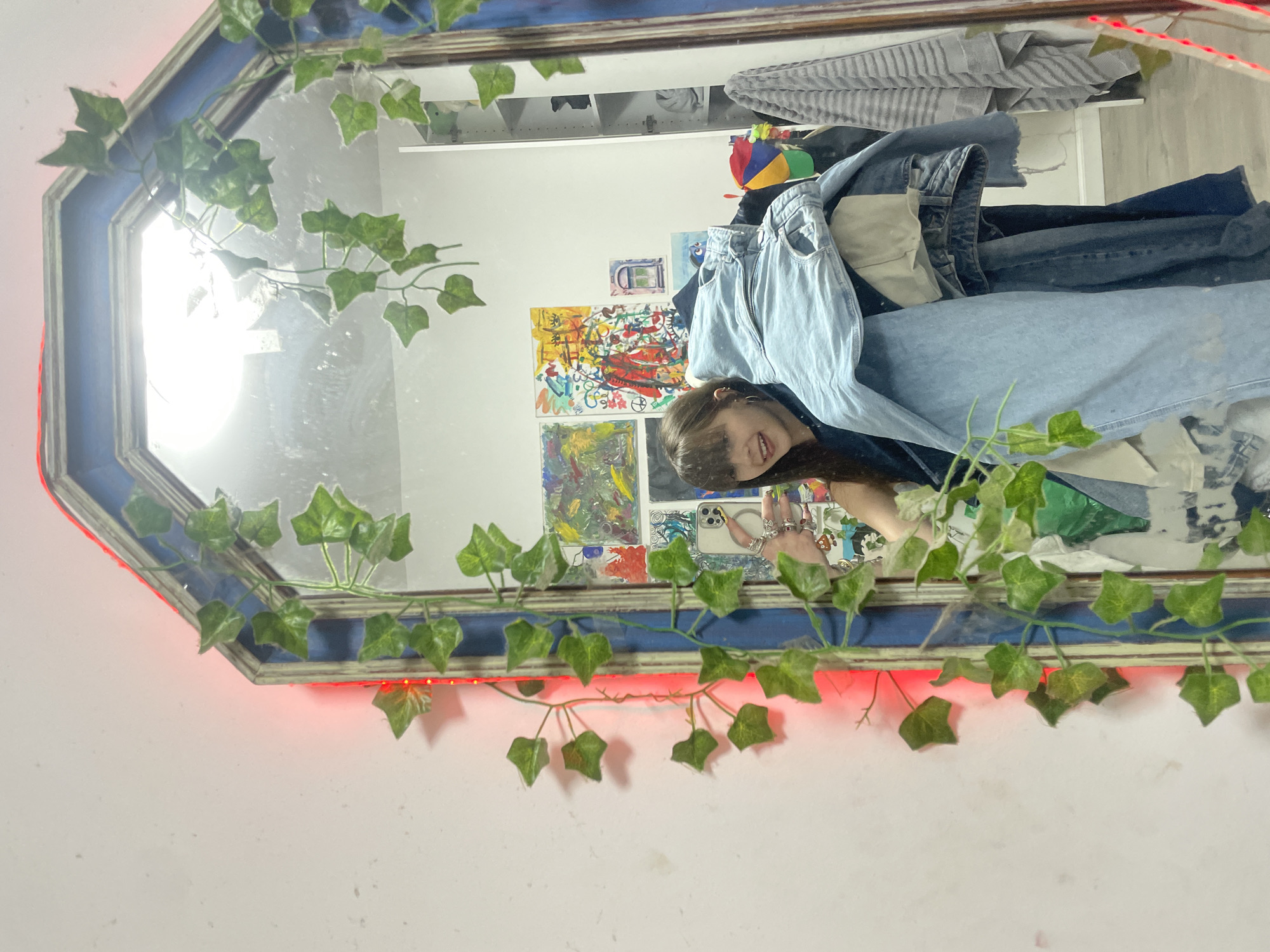 The image shows a selfie taken in a mirror decorated with artificial ivy and red LED strip lights. The mirror's frame is octagonal and painted blue. The reflection includes a young woman smiling, holding a smartphone with multiple rings on her fingers, along with various items including folded and hanging clothing, artwork on a wall, and a shelf with miscellaneous items. The background suggests an interior space, likely a bedroom. It is not possible to determine the location in Raanana, Israel, from the image.