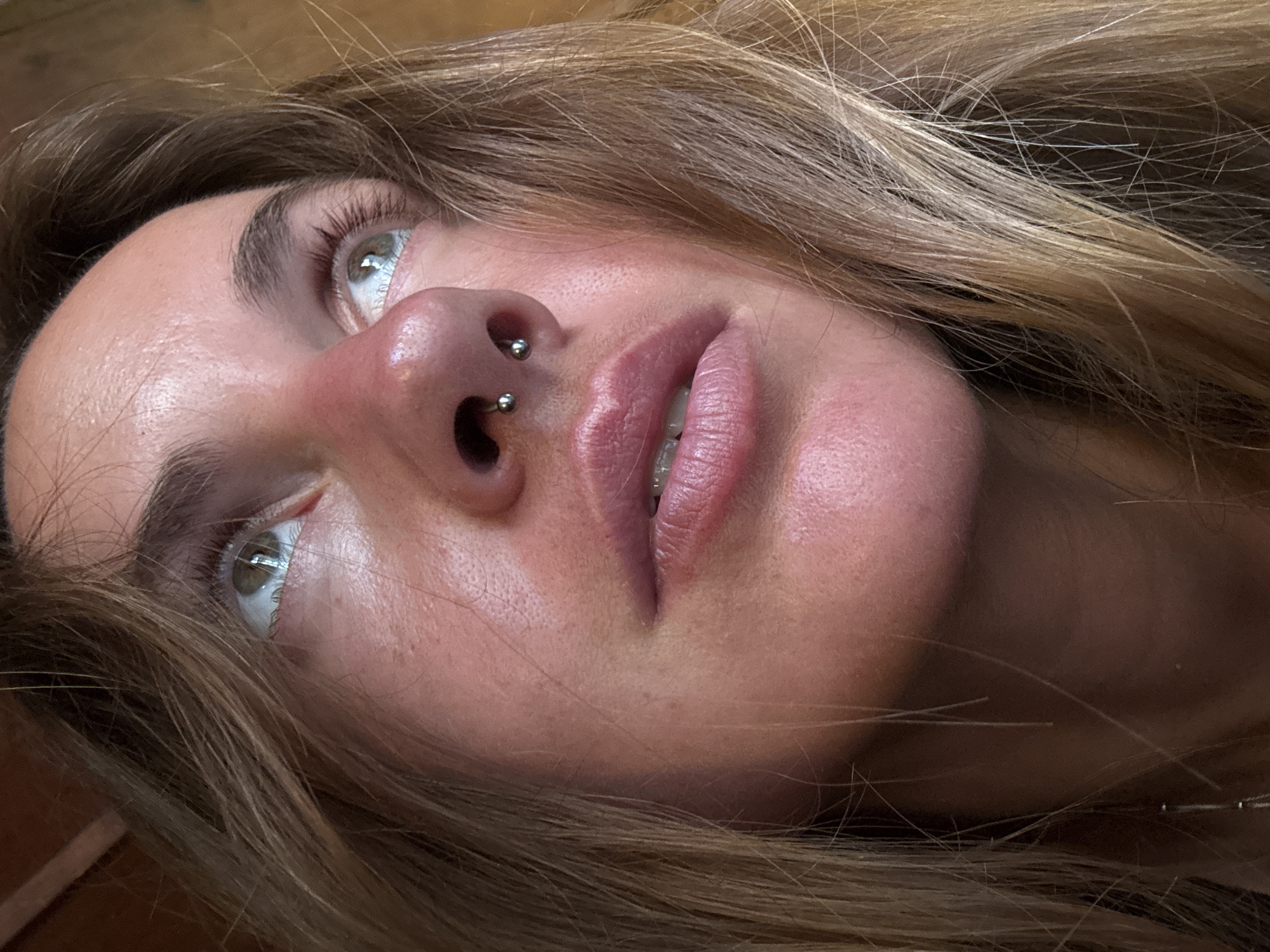 A close-up, low-angle, indoor shot shows a fair-skinned young woman with blonde hair and green eyes looking upwards and to her right, with her head tilted back slightly. Her nose has a septum piercing. Her lips are parted, revealing dental braces. Her face is flushed, and her skin appears to have texture from pores and some blemishes. Her hair is light brown and blonde, with strands falling across her face and shoulders. The background is out of focus, appearing to be wooden. The lighting is warm and directional, creating highlights on her skin and hair.