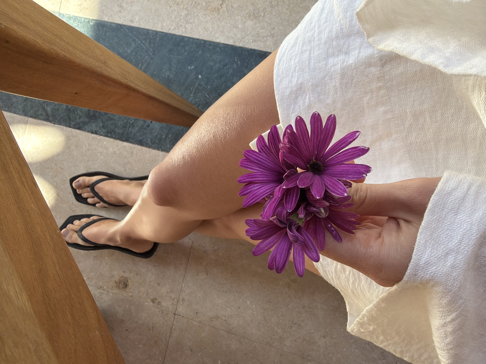 A high-angle shot captures a person relaxing in a sunlit setting, likely at a resort in Sharm El Sheikh, Egypt. The main subject is the person's bare legs, crossed at the knees, with their feet wearing simple black flip-flops. A hand gently holds a small cluster of vibrant purple daisy-like flowers, possibly African Daisies (Osteospermum), near one knee.
The person is clothed in a loose-fitting, light-colored, textured fabric, resembling a white or cream dress or robe, which drapes softly around their body. Bright, direct sunlight illuminates the scene, casting strong, distinct shadows on the floor and highlighting the texture of the skin and fabric. This lighting suggests either a morning or late afternoon setting. The flooring consists of large, light beige or grey tiles, with a contrasting strip of darker blue or grey tiles visible under a wooden structure, likely a chair or table leg, in the upper left of the frame. The overall impression is one of peaceful leisure and natural beauty. No visible text is present in the image.