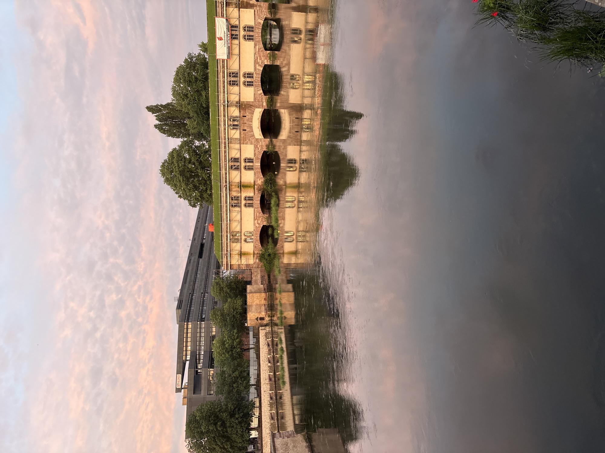 This image captures a wide shot of the Ill river in Strasbourg, France, featuring the historic Barrage Vauban and adjacent structures, with a modern building in the background under a pastel-colored sky.
The sky occupies the upper third of the frame, displaying scattered cirrocumulus clouds tinged with soft pink and light blue hues, suggesting either sunrise or sunset. The water below is calm and dark, reflecting the sky and the structures along its banks with notable clarity.
In the midground, the Barrage Vauban, a substantial stone structure, spans the river. It features multiple arched openings at water level and a series of rectangular windows with arched tops above, indicating a covered walkway or internal space. The structure's facade is light brown and beige, partially obscured by extensive scaffolding, which suggests ongoing maintenance or renovation. A white banner with red text, displaying "schoenenberger" and a phone number (03 88 79 19 21), is affixed to the scaffolding on the right side of the barrage. The roof of the barrage is covered with green grass or vegetation. To the immediate left of the main barrage, a smaller, square stone structure with an arched opening, characteristic of the Ponts Couverts complex, is visible.
Along the riverbanks, scattered green trees with dense foliage are present, particularly to the left of the barrage. In the far background, behind these trees, a large, dark-grey, multi-story modern building is prominent. This building features numerous horizontal windows and a distinct rooftop structure. A sign on its roof clearly displays the word "ALSACE" with a logo.
In the lower right corner, a cluster of green reeds and a few small red flowers emerge from the water. No individuals are visible in the scene.