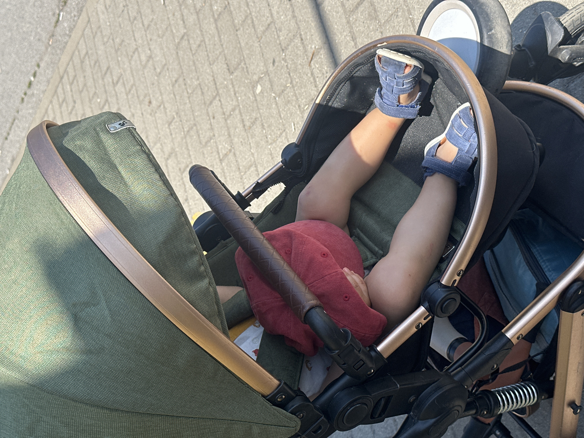 A close-up, medium shot captures a child's legs and feet sticking out of a stroller. The child is wearing blue sandals and has a red cap over their head. The stroller is green with gold accents. The background shows a paved sidewalk and a road, suggesting an outdoor urban setting. The lighting indicates it is daytime, with shadows cast by the stroller and a person holding it. The scene is likely in Sofia, Bulgaria, given the context of the request.