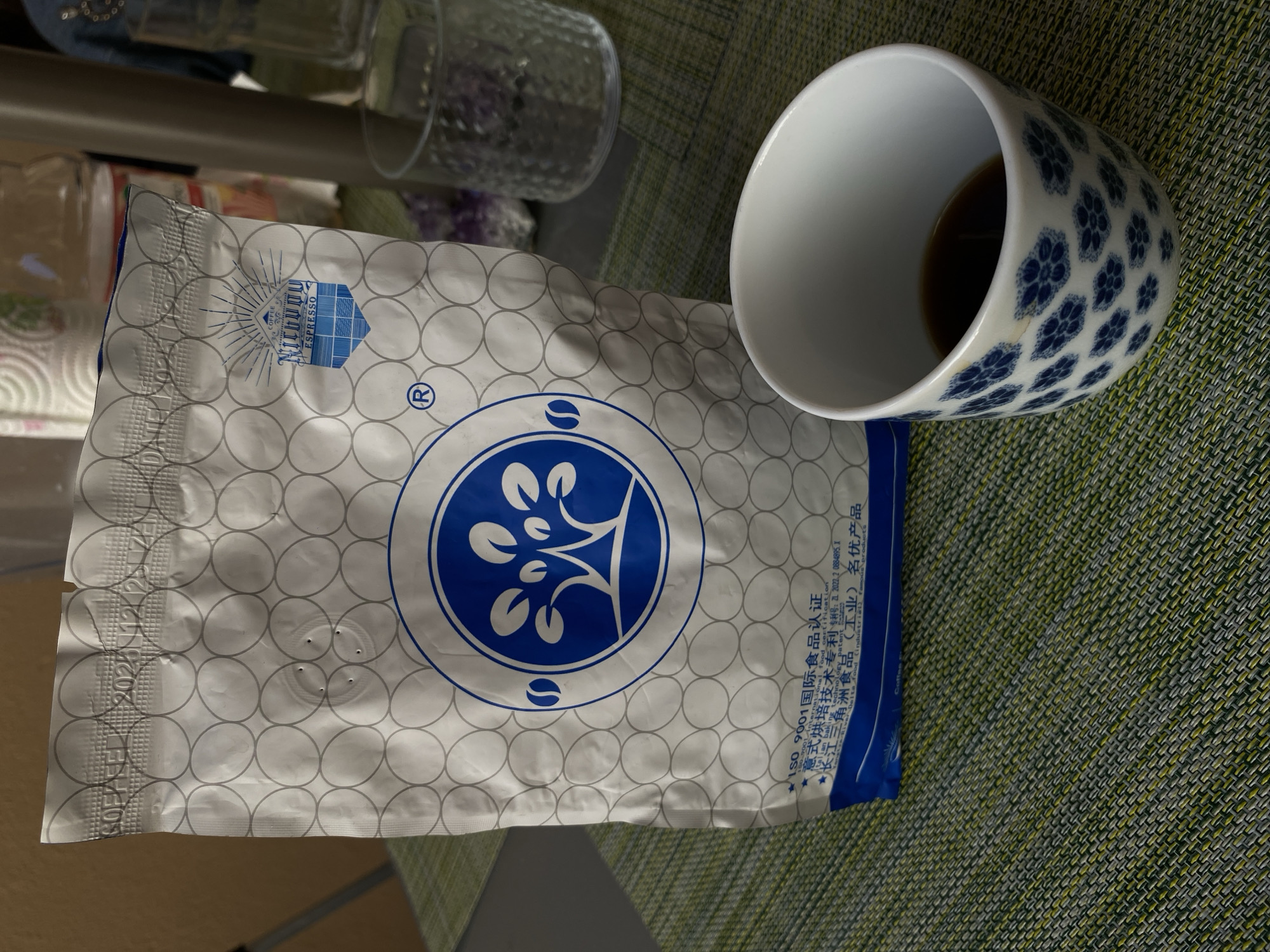 The image shows a partially filled teacup with blue floral patterns next to a bag of espresso coffee on a textured green surface. The coffee bag is white with a repeating pattern of gray circles. It features a large blue logo in the center with a tree-like design and smaller blue logos near the top and bottom. Text in Chinese and English is printed near the bottom right of the bag, including "ISO 9001 international certification" and references to Italian roasting technology. The scene is indoors, likely on a table.