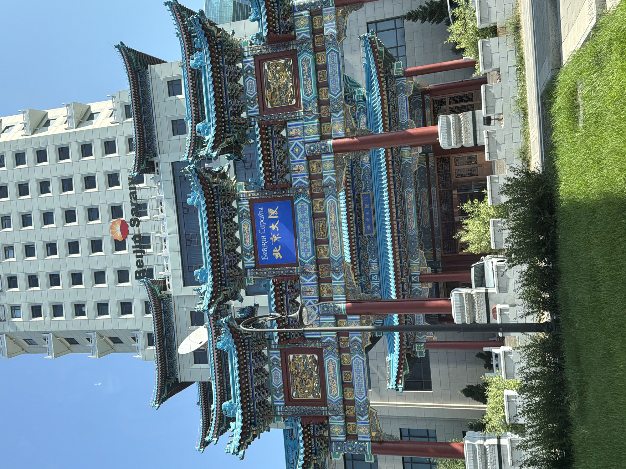 A multi-tiered traditional Chinese-style gate with ornate blue and gold detailing stands prominently in front of a modern, beige high-rise building under a clear blue sky. The gate features multiple roofs with upturned eaves, red columns, and intricate carvings. A blue sign with Chinese characters is visible on the gate. The high-rise building has numerous windows and balconies. In the foreground, there is a patch of green grass, a sidewalk, and a low, decorative white stone wall with bushes and trees. A street lamp is positioned to the left of the gate. A white car is partially visible behind the wall. This is the "Beijing Palace" hotel in Astana, Kazakhstan, a landmark representing Sino-Kazakh relations.