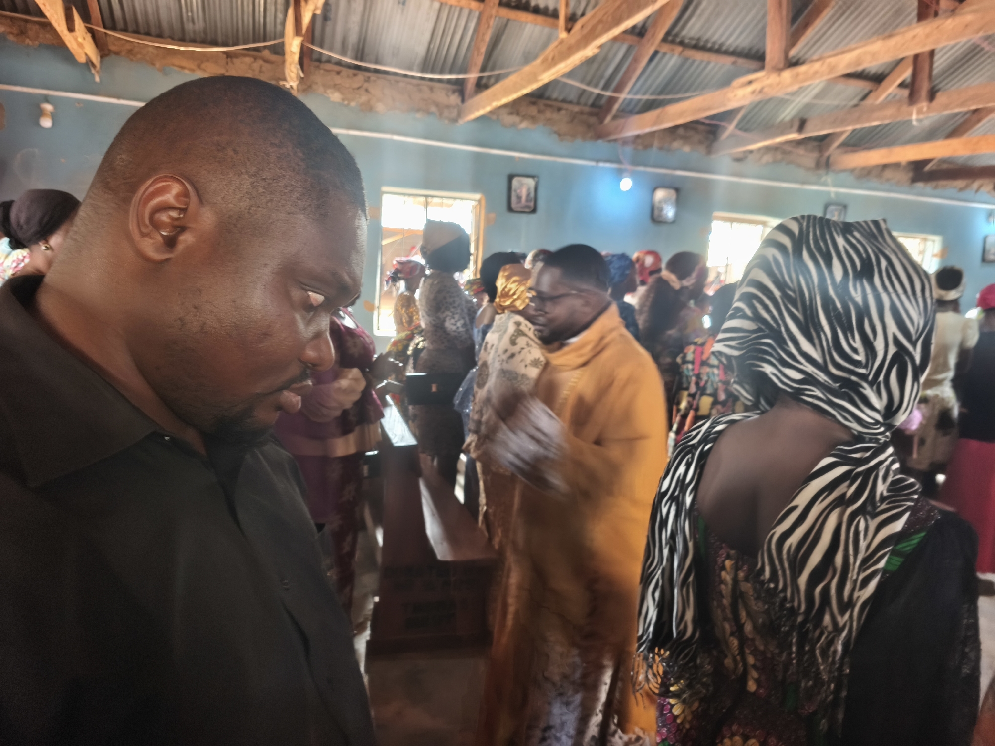The image depicts the interior of a room, possibly a church or community hall, in Nabatum, Nigeria. Multiple individuals are present, most of whom appear to be standing. In the foreground, a man with short hair is visible from the side, wearing a dark shirt. Another person is seen from behind, wearing a zebra-striped head covering. In the background, other individuals are gathered, some wearing glasses and colorful clothing. The room has a light blue wall color and exposed wooden beams supporting the ceiling. There are framed pictures on the wall and windows that let in natural light.