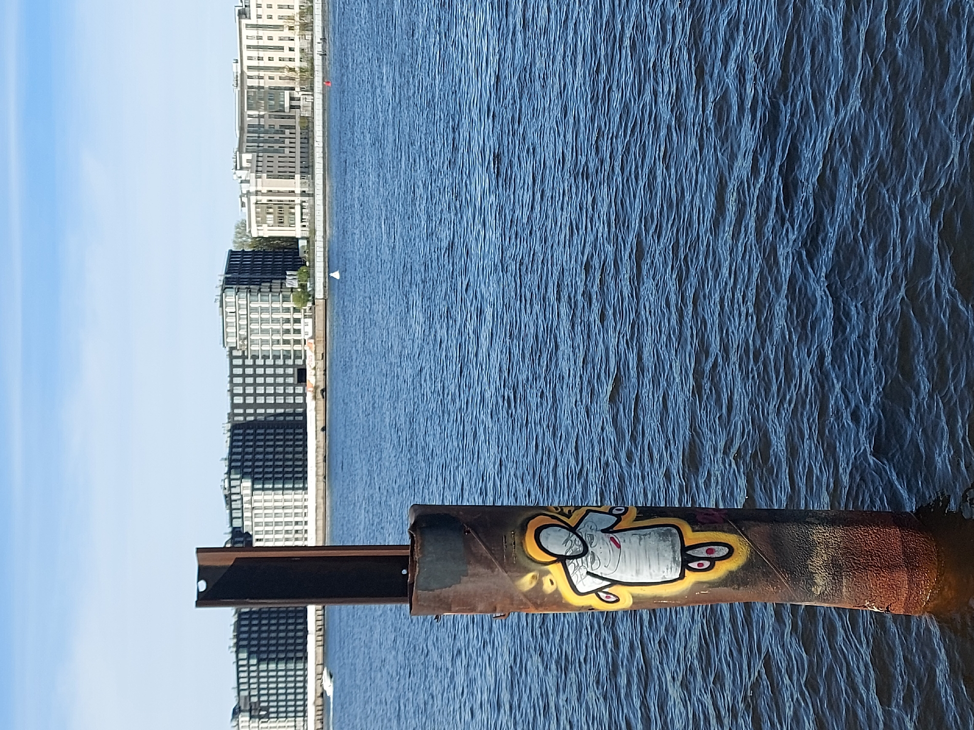 Here is a description of the image:
The image shows a view of the Neva River in Saint Petersburg, Russia. The river is wide and choppy with visible waves, occupying a significant portion of the frame. The opposite bank is lined with modern apartment buildings. A metal pipe with graffiti sits in the foreground. The graffiti is of a cartoonish figure with yellow and red accents. A brown beam extends from the pipe toward the buildings. The sky is partially visible and blue with some clouds.