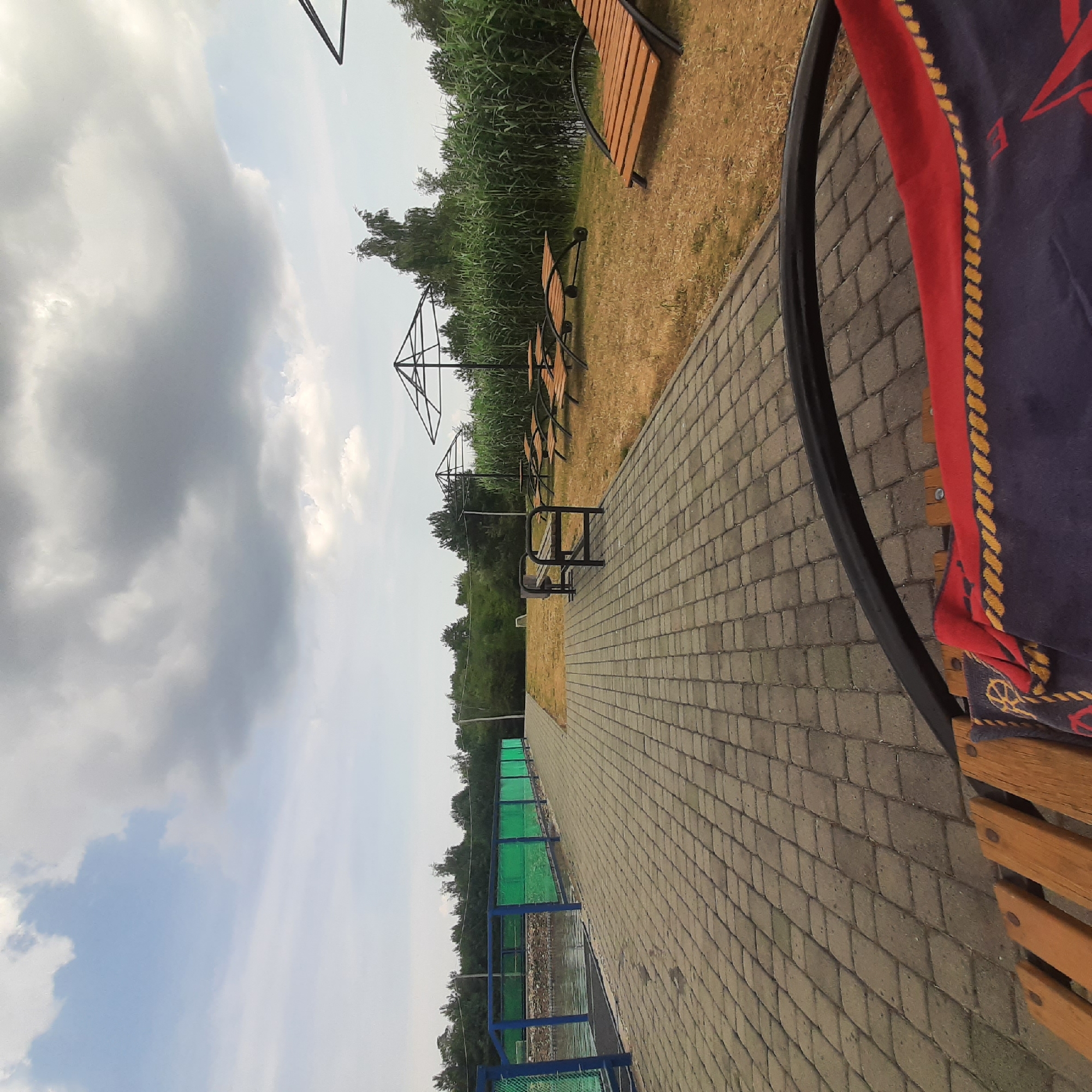 This is a wide-angle, eye-level shot of an outdoor recreational area. A paved walkway, made of gray interlocking stones, occupies the lower left and foreground of the image. To the left of the walkway is a body of water, bordered by a blue and green fence. Beyond the fence and water, lush green trees form a dense treeline.
The walkway leads to an area with sparse, dry grass and several amenities. There is a dark metal bench with a trash receptacle next to it. Further back, three wooden lounge chairs with black metal frames are arranged on the grass. Two large, black, umbrella-like structures, likely shade structures or canopies, are also present. Behind these structures, tall, reedy plants grow densely, bordering the water. In the distance, more trees are visible against a cloudy sky. The overall impression is of a somewhat unkempt but functional outdoor leisure space. The image does not contain any discernable human presence. The location is identified as Dubrivka, Ukraine.