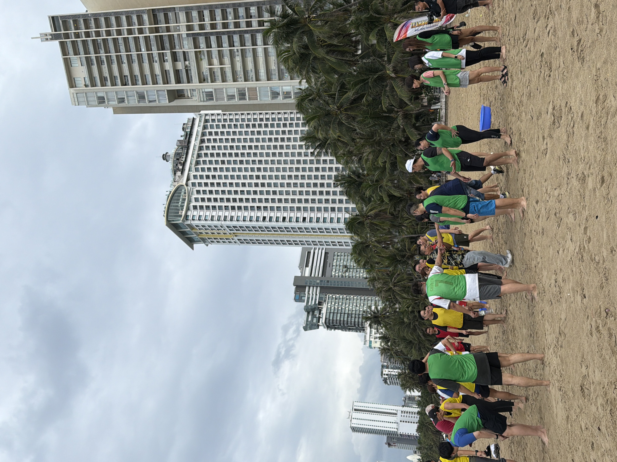 This is an eye-level, medium shot photograph taken on a sandy beach in Nha Trang, Vietnam, with a cloudy sky overhead. In the background, several tall, modern buildings are visible, including a prominent white building with many windows and a yellow-capped top, and another taller, beige building with balconies. Lush palm trees line the edge of the beach between the people and the buildings.
In the foreground, a group of approximately 20 to 25 people are gathered on the sand, appearing to be participating in a team-building activity or informal game. Many of the individuals are wearing matching brightly colored vests, predominantly green and yellow, over casual clothing. Some are wearing athletic shorts and t-shirts, while others are in longer pants. The people are in various poses, some standing and talking, others bending down or looking towards each other, suggesting active engagement in a shared activity. One person in a yellow vest is holding a blue plastic tub. A rolled-up banner with text is visible on the right side of the frame. The overall atmosphere appears to be relaxed and social, with people barefoot on the sandy shore.