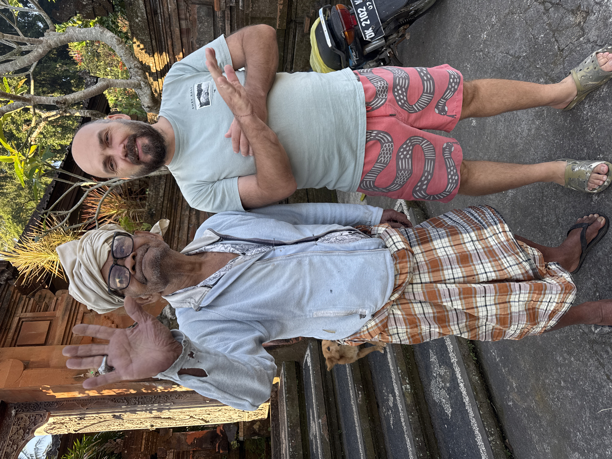 The image shows two men standing in front of a stone structure in Tampaksiring, Indonesia. The man on the left is older, wearing a light blue jacket over a patterned shirt and a plaid sarong. He has a turban on his head and glasses, and he is waving with his right hand. The man on the right is younger, with a bald head and beard, wearing a light blue t-shirt and pink shorts with a snake pattern. He has his arms crossed. A small brown dog is visible on the steps behind the older man. A motorcycle with a yellow cover is parked to the right of the younger man. The environment appears to be outdoors, with trees and foliage visible in the background, and the architectural style suggests a temple or traditional building. The overall lighting suggests it is daytime.