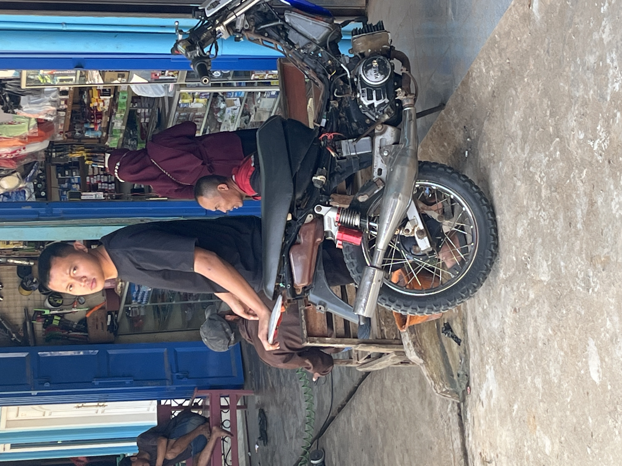 The image captures a street scene in Karang Sari, Indonesia, focusing on a motorcycle parked in front of a shop. In the foreground, a partially disassembled motorcycle with its engine exposed is the primary subject. A young man in a black t-shirt is standing near the motorcycle, looking towards the camera. Behind him, a woman in a maroon hijab is browsing inside the shop, which is filled with shelves of merchandise. Another man, wearing a red and black shirt, is seated on a wooden bench near the motorcycle, engaged in conversation. Further to the left, another individual is seated on a reddish bench. The environment appears to be a bustling commercial area with concrete flooring and building facades.