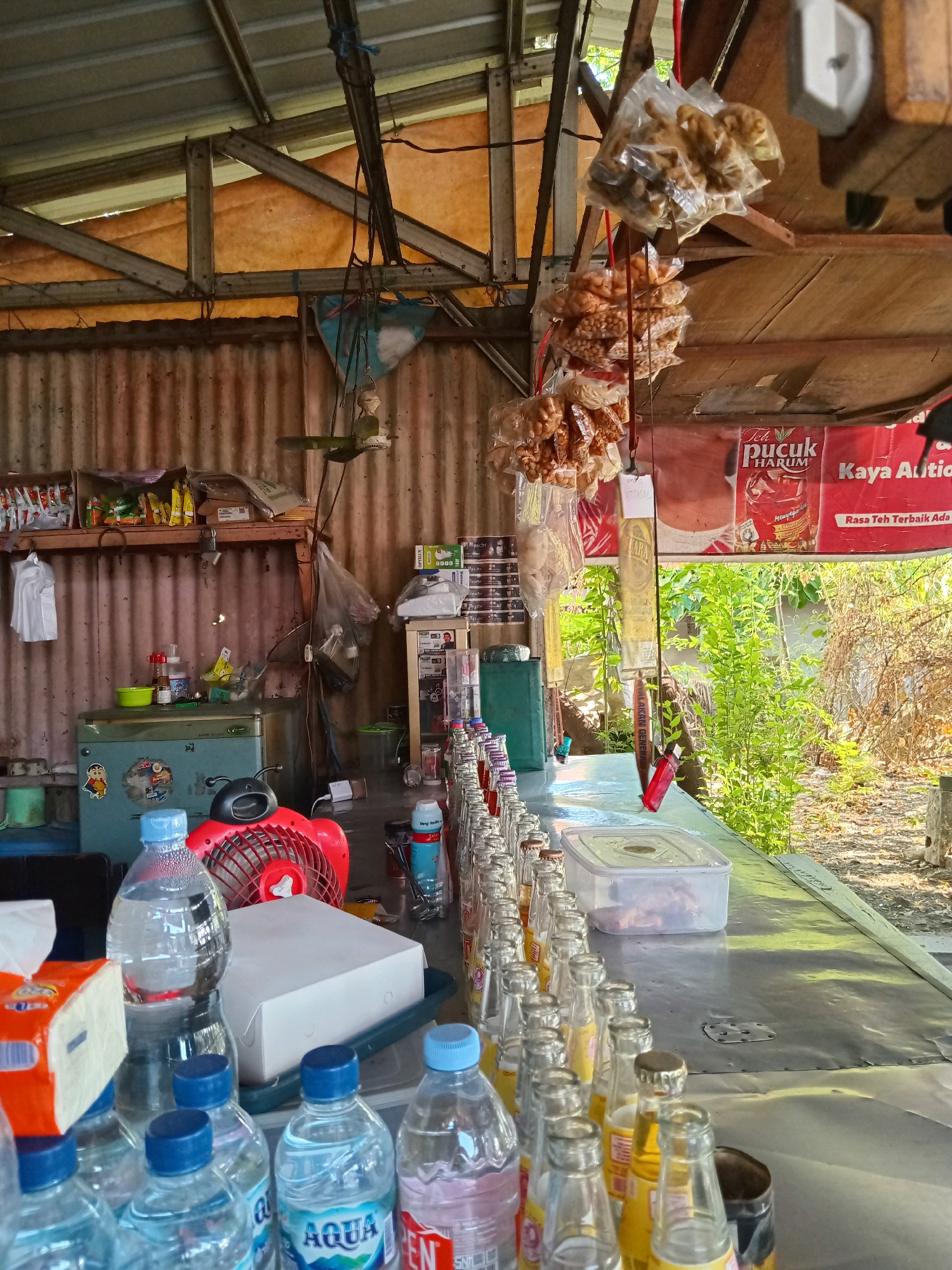 The image depicts the interior of a small, open-air food and beverage stall in Kembangan, Indonesia. The stall is constructed with corrugated metal walls and a metal roof supported by wooden beams and metal framing. A long, gray counter dominates the foreground, laden with rows of bottled drinks. In the background, shelves hold various packaged goods, including snacks and boxes. To the left, a turquoise refrigerator is visible, adorned with stickers. A red electric fan is positioned on the counter. Hanging from the ceiling are plastic bags filled with snacks, and a red banner with Indonesian text, likely advertising beverages, is displayed. Outside the stall, lush green foliage is visible, suggesting an outdoor setting. The overall impression is one of a modest, functional establishment catering to local customers.