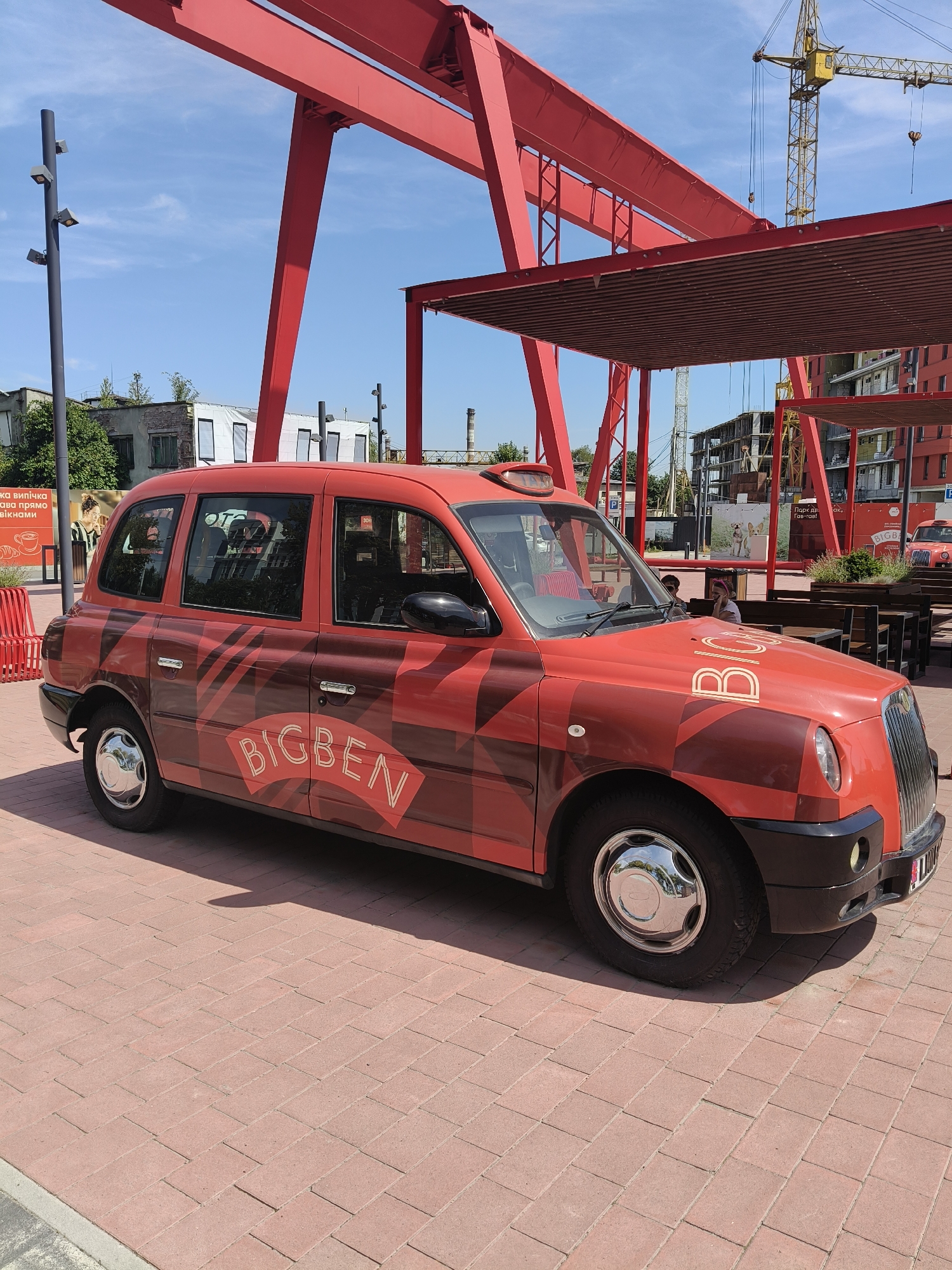 A bright red, classic-style taxi with "BIGBEN" branding is parked on a pedestrian walkway made of red brick pavers. The taxi has a black roof, black trim, and chrome hubcaps. The vehicle is adorned with a pattern of darker red diagonal stripes and a large "BIGBEN" logo on its side and front hood. In the background, a large red industrial-style structure looms overhead. Further back, construction cranes and unfinished buildings suggest an urban development area. A clear blue sky with some wispy clouds serves as the backdrop. The scene appears to be outdoors in an urban setting, possibly a public square or a newly developed district. The specific location of Lviv, Ukraine, is not discernible from the image itself.
