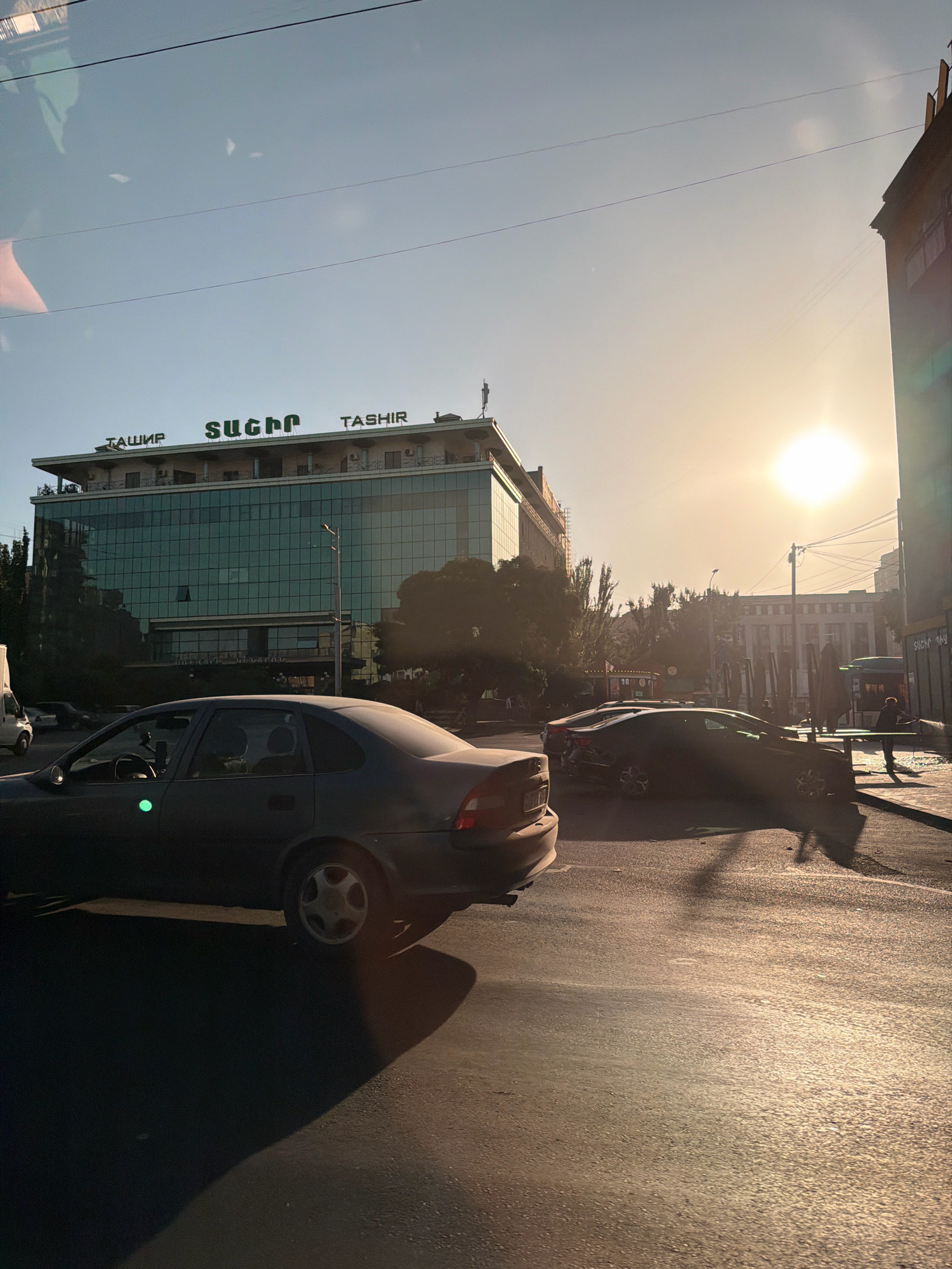 The image shows a street scene in Yerevan, Armenia, during daylight hours. The sun is visible in the sky, casting long shadows from the vehicles and buildings. In the foreground, a dark-colored sedan is parked on the right side of the street, its front passenger side showing a green reflective sticker. Across the street, a modern glass-fronted building with the name "TASHIR" and "ТАШИР" in green lettering is prominent. Several other vehicles are visible on the street, including a dark sedan further down the road and a white van on the left. The street is paved and appears to be an urban environment with some trees and other buildings in the background. A person is partially visible on the sidewalk to the right, holding what appears to be a hose. The overall atmosphere suggests a sunny day with moderate traffic.
