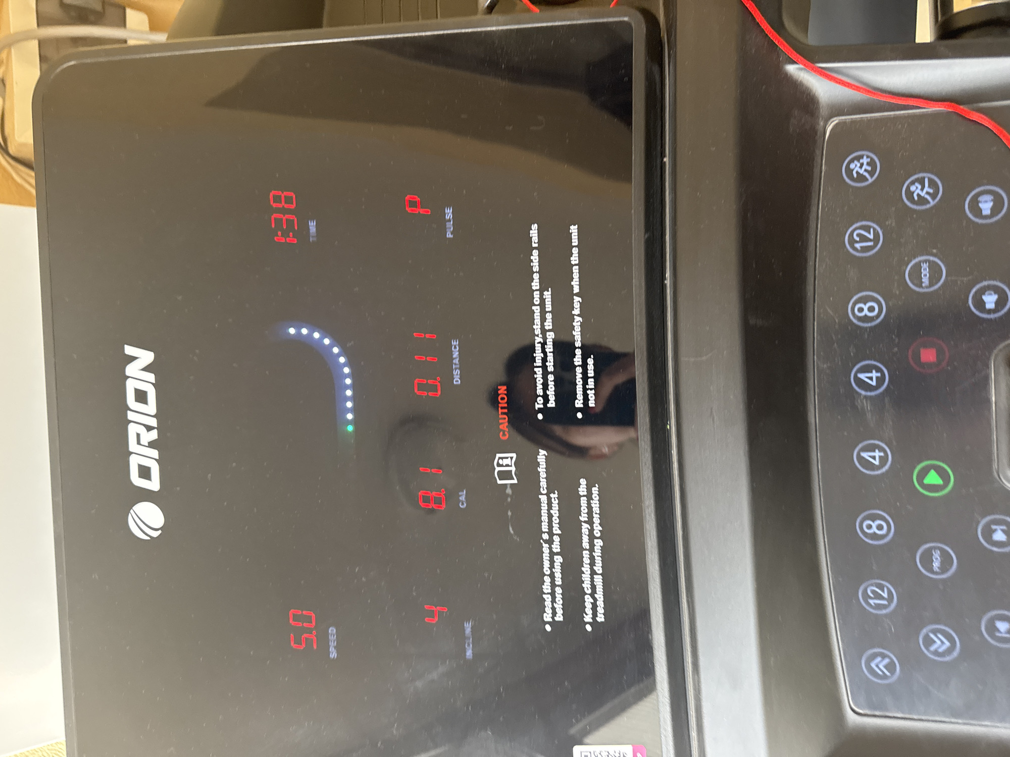 A close-up shot of an Orion treadmill console. The display shows: Time 1:38, Distance 0.11, Calories 8.1, Speed 5.0, Incline 4 and Pulse P. Cautionary notes are present regarding safety procedures. To the right, there are various buttons on the console: 12, 8, 4, Mode, Prog, and arrow buttons. The location is unknown and could not be identified based on the image.
