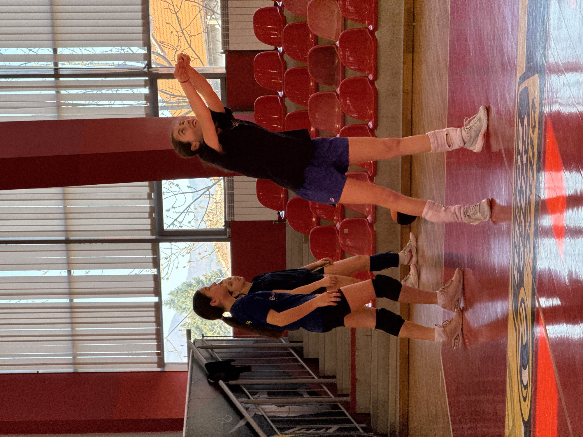 Two young women are in a gymnasium in Etropole, Bulgaria. The gymnasium has red plastic seats along one wall, large windows with closed blinds, and a polished red wooden floor marked with patterns. The woman in the foreground is wearing a black t-shirt, blue shorts, white socks and shoes, and is stretching her arms overhead. The woman in the background is wearing a blue shirt, black shorts and knee pads, and tan colored shoes. She is also stretching. A set of aluminum bleacher seats is visible at the bottom left.