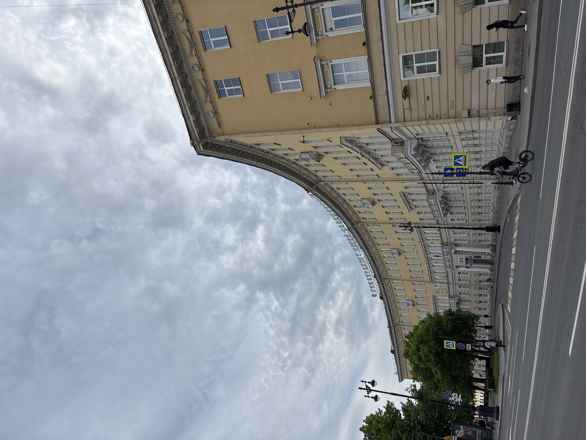 The image, oriented correctly, depicts a wide urban street scene in Saint Petersburg, Russia, under an overcast sky. A grand, curved building with a light yellow facade and numerous rectangular windows dominates the left side of the frame, extending from the foreground into the midground. The building exhibits classical architectural details, including decorative cornices and window frames.
In the foreground, on the right side of the street, a person is standing on a BMX bicycle, seemingly performing a trick or posing, with their back to the viewer. Further down the sidewalk on the right, two other individuals are walking. The street itself is multi-laned, with clear white road markings, including double solid lines and pedestrian crossing stripes. Several streetlights are visible, along with lush green trees lining parts of the sidewalk and a small median.
Visible street furniture includes a prominent blue and yellow pedestrian crossing sign, and a blue directional sign with a white arrow. On the curved building, the Cyrillic letters "ДЛТ" (DLT) are visible, indicating the name of a department store. The sky is filled with varying shades of grey clouds, suggesting a cloudy but bright daytime.