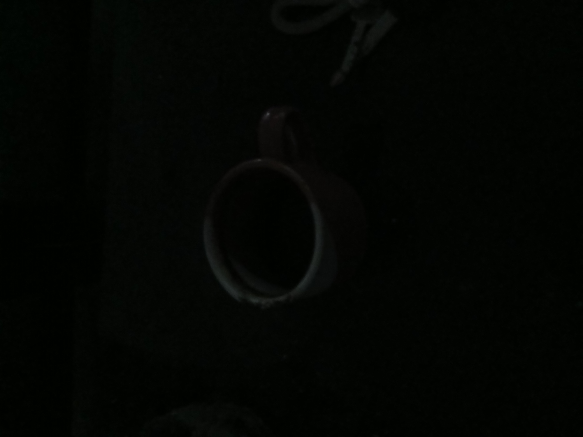 A dark, low-light image shows a red mug filled with a dark liquid, possibly coffee, positioned on a dark surface. To the right of the mug, a pair of scissors is visible. The environment appears to be an indoor setting, possibly a desk or table, given the presence of these objects. The overall lighting is poor, obscuring many details of the surroundings. There are no people or animals visible in the frame. No specific location in Kademangan Kulon, Indonesia, can be identified from the visual information provided in the image.