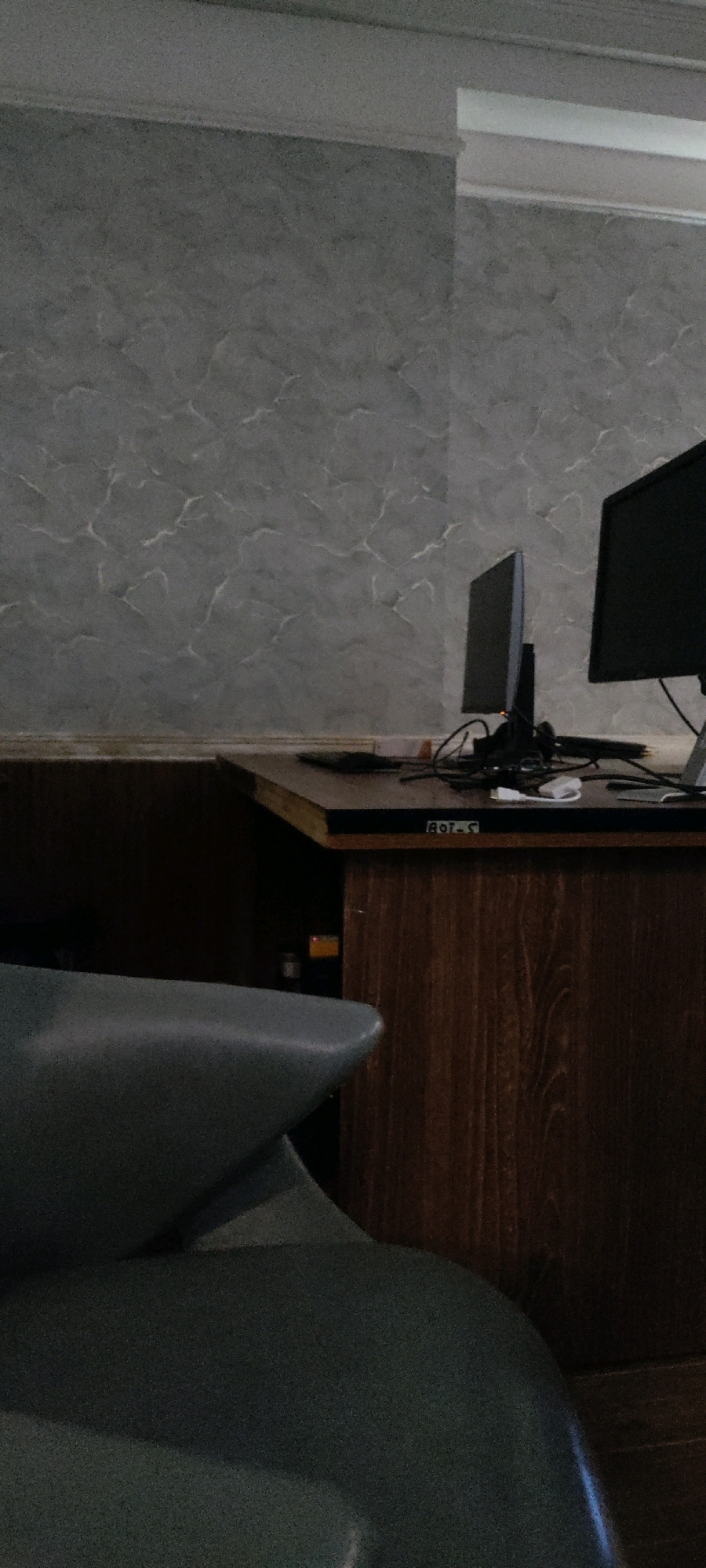 The image depicts an indoor scene with a desk and two computer monitors. The desk is made of wood and has a brown finish. On the desk, there are cables and a white object, possibly a USB drive. One monitor is black and is positioned on the right side of the desk. The other monitor is silver and is located to the left of the black monitor. The wall behind the desk has a textured grey finish with a marble-like pattern. In the foreground, a portion of a dark grey chair is visible. The scene appears to be a workspace or an office. The location is identified as Khana Dak, Pakistan.