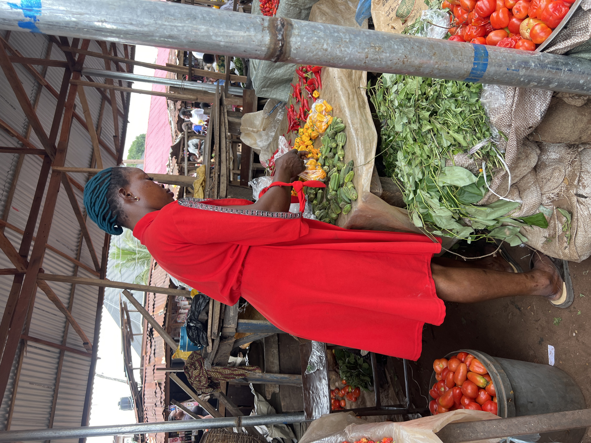 A woman in a bright red dress stands at a market stall in Nkpor, Nigeria, arranging produce. Her teal-colored hair is braided and pulled back. In front of her, on a beige-covered table, are piles of green okra, yellow peppers in plastic bags, and red chili peppers. Behind her, a large black bucket overflows with red tomatoes. The market has a rustic setting with wooden structures and corrugated metal roofing. Other market-goers are visible in the background, suggesting a busy commercial area. The ground is dirt, typical of an outdoor market.