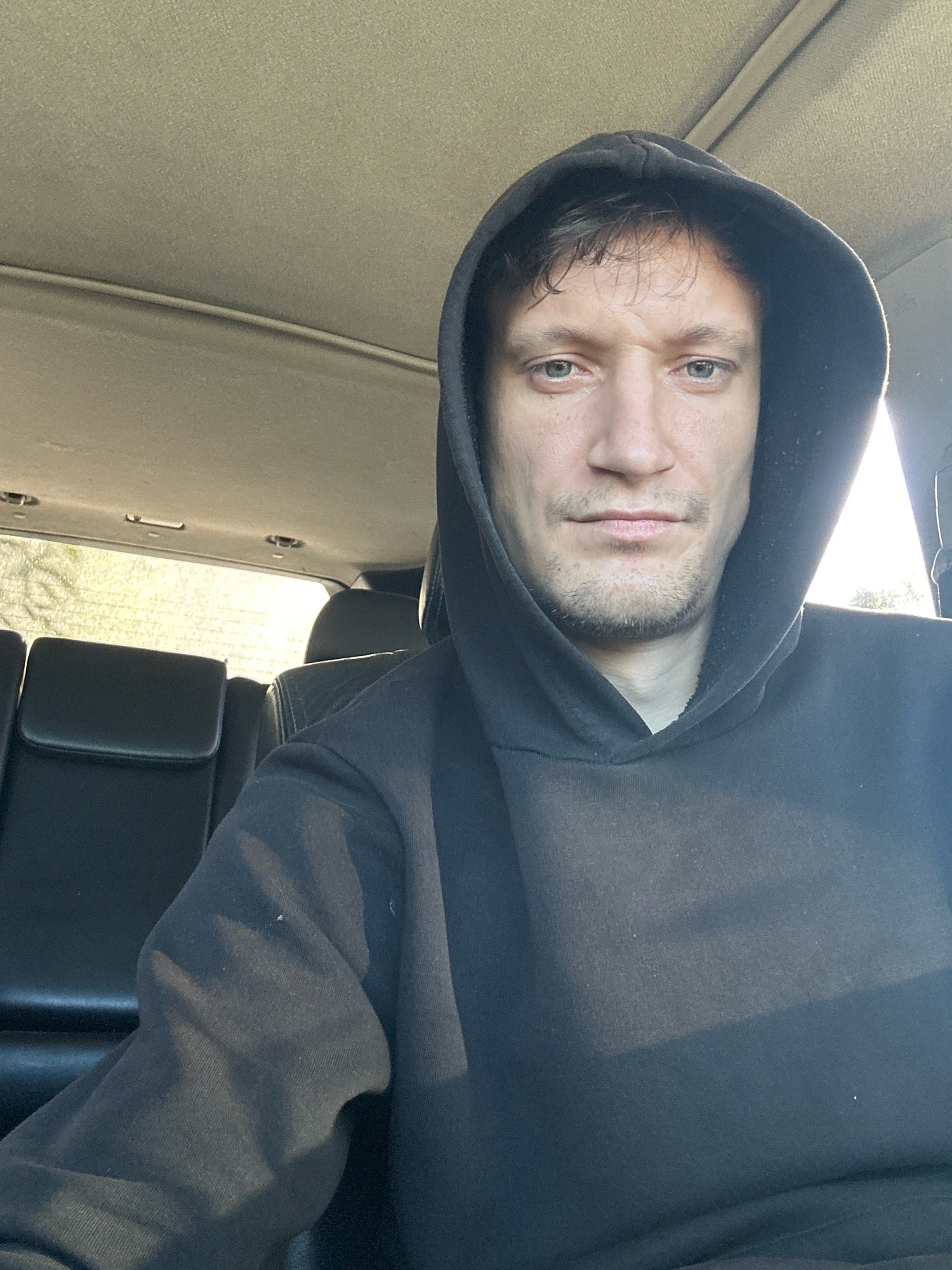 A man wearing a black hooded sweatshirt is seated in the front passenger seat of a vehicle. The vehicle appears to be a sport utility vehicle, with black leather seats visible in the background. The man is looking directly at the camera and has short, dark hair. Sunlight is streaming in from the right side of the frame, casting shadows on the man's face and hood. The interior of the vehicle is dimly lit, with the ceiling of the car visible above. The scene is consistent with a selfie taken inside a vehicle. There is no information to suggest this location is in Makiv, Ukraine.