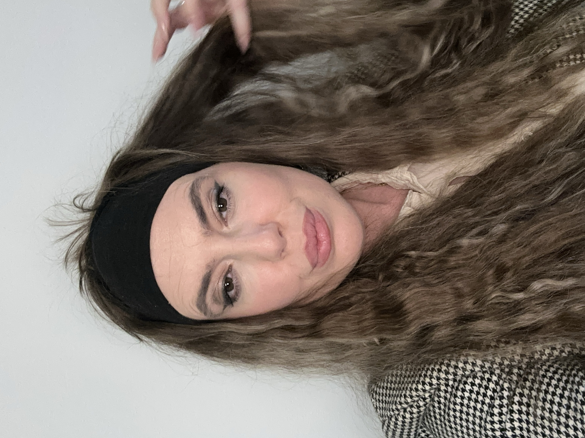A woman with light skin and brown eyes, wearing a black headband, is looking at the camera. She has long, wavy, light brown hair. She is wearing a light-colored top and a black and white houndstooth patterned jacket. A fingernail is visible in the upper left of the image. The background appears to be a plain, white wall. There is no information provided that indicates the location is Tbilisi, Georgia.
