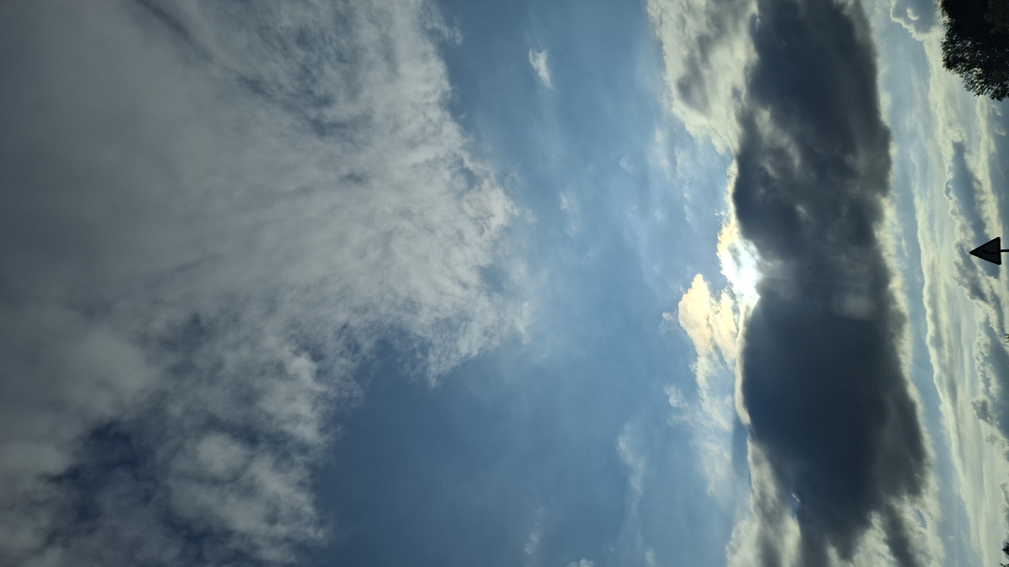 This media file shows a cloudy sky in Забегалово, Russia. The clouds are a mix of dark, heavy formations and wispy, lighter ones, indicating varied weather conditions. The sun is partially visible, peeking through a gap in the clouds, casting rays of light that highlight the textures and depth of the sky. Some of the lighter clouds appear to be streaks, possibly indicating wind. The overall scene suggests it is daytime, likely late afternoon or early evening given the angle of the sun and the contrast between the bright sky and the darker clouds. There are no visible people or specific objects, aside from a small, dark triangular sign on a pole on the right side of the image, which is partially obscured by clouds and appears to be a directional marker. The top right edge of the image shows the dark silhouette of trees, suggesting a natural or semi-rural setting. The lighting creates a dramatic and somewhat moody atmosphere. No text is visible in the image.