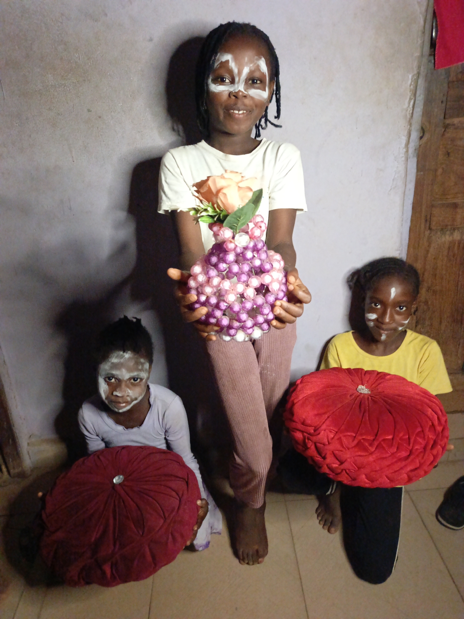 Three barefoot young girls stand in a room. The girl in the center is taller and holds a beaded ball with artificial roses. All three girls have white face paint applied. The two girls on either side of the taller girl each hold a red velvet cushion. The room is simply furnished, with a grey wall and a tiled floor. There is a partially visible wooden door in the background. The location is Unknown, Unknown.